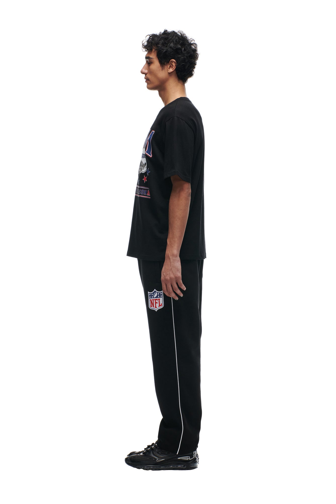 A man stands in profile against a white background, wearing the 6PM NFL VS 6PM T-SHIRT BLACK, black NFL sweatpants with white piping, and black sneakers.