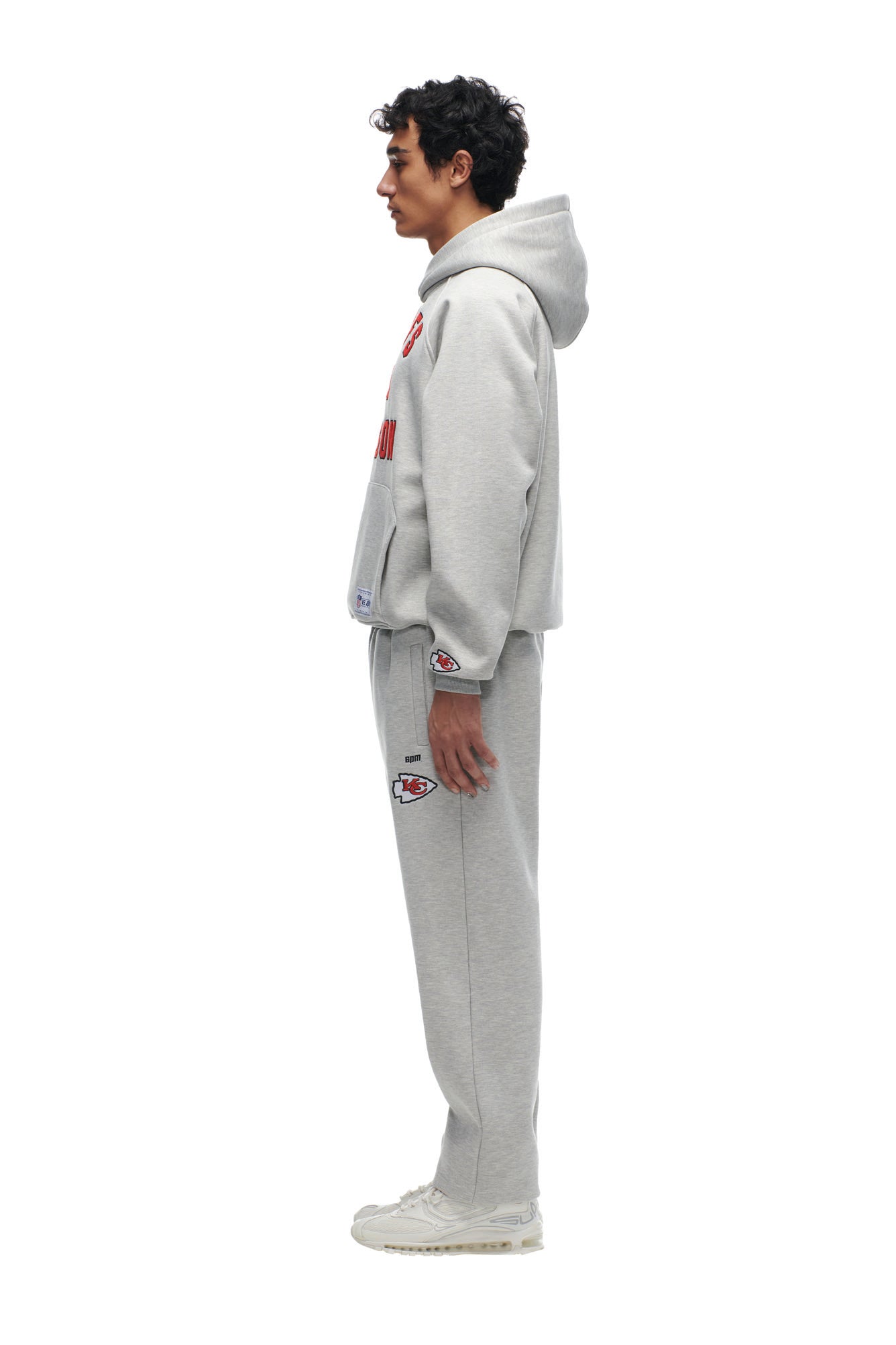 A person stands in profile against a plain white background, wearing the 6PM CHIEFS SEASON HOODIE and matching sweatpants with red and black logos, paired with white sneakers.