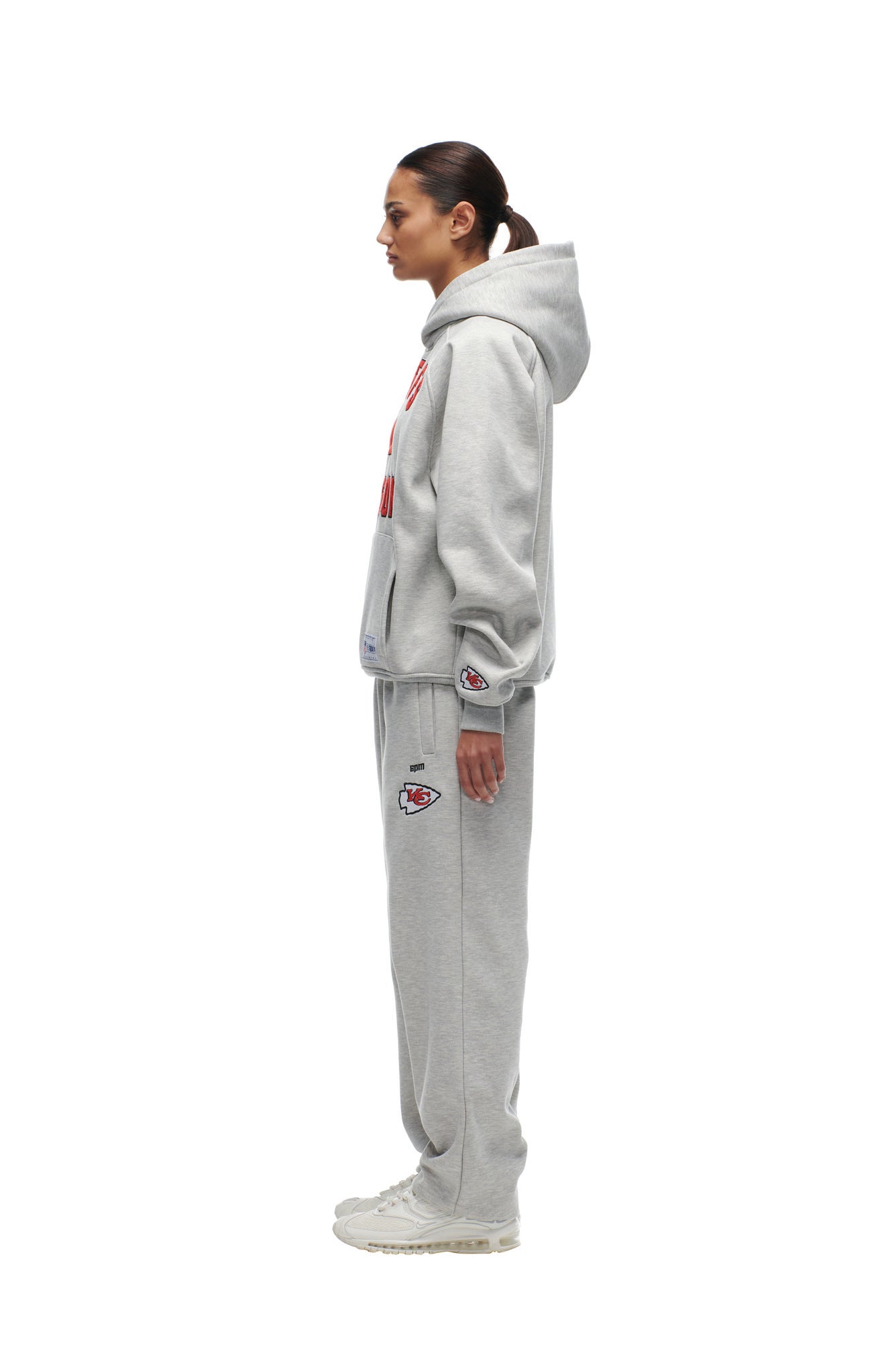 A woman stands in profile against a white background, wearing the 6PM CHIEFS SEASON HOODIE and matching sweatpants with red and white logos, paired with light-colored sneakers.