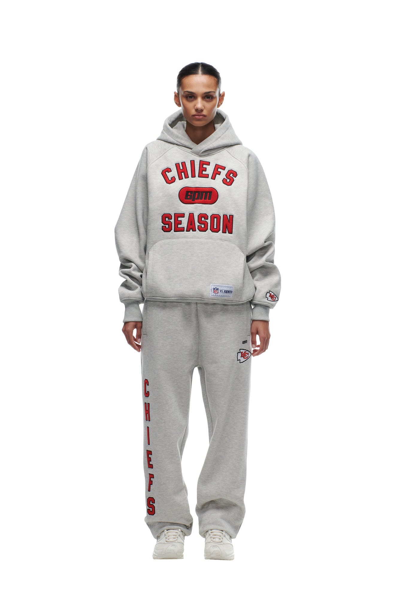 A person faces forward in a gray 6PM CHIEFS SEASON HOODIE and matching sweatpants with red and black text and logos, paired with white sneakers, against a plain white background.