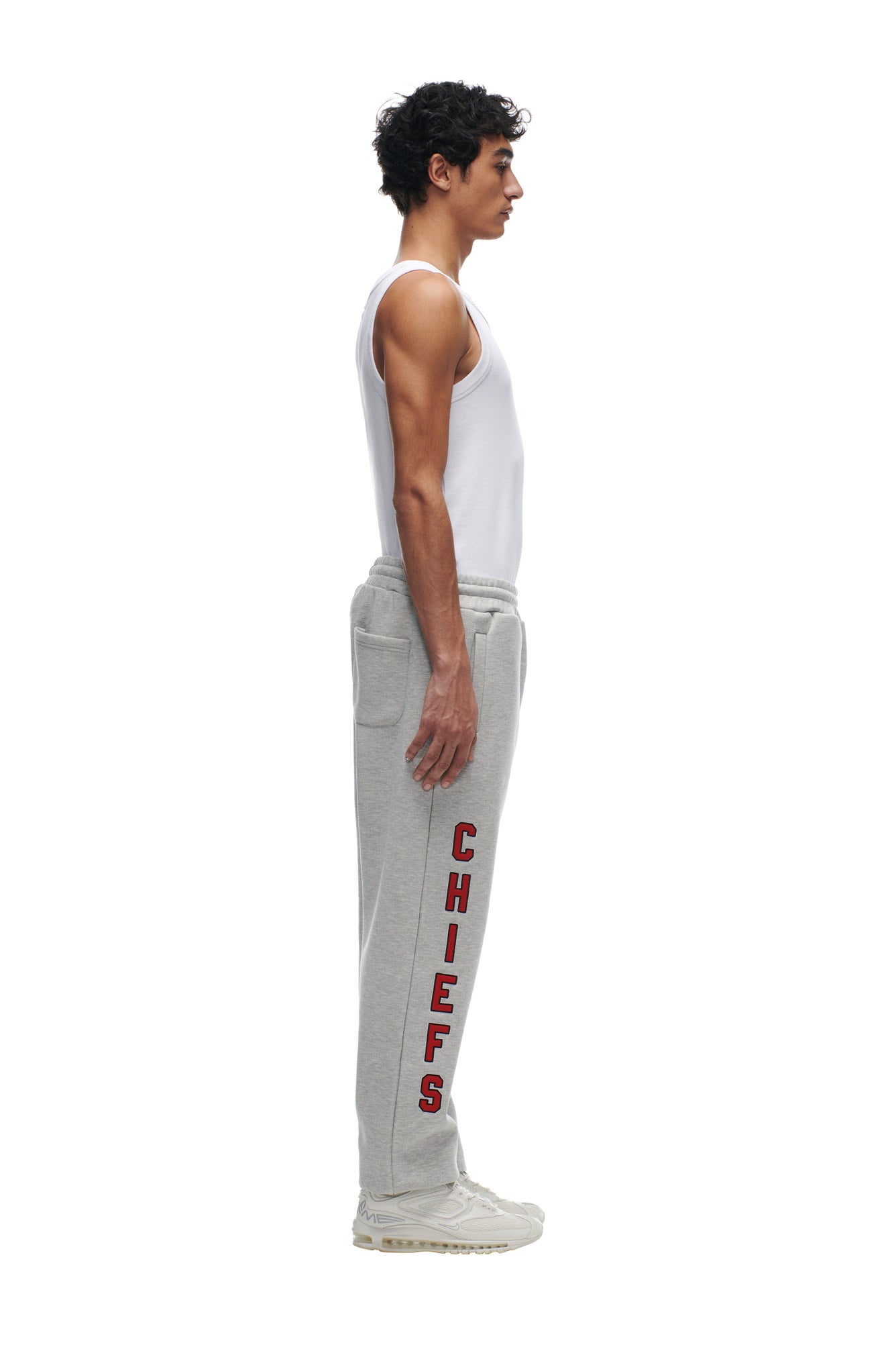 A man wearing a white tank top and 6PM CHIEFS SEASON JOGGER sweatpants with red "CHIEFS" lettering stands in profile against a white background.