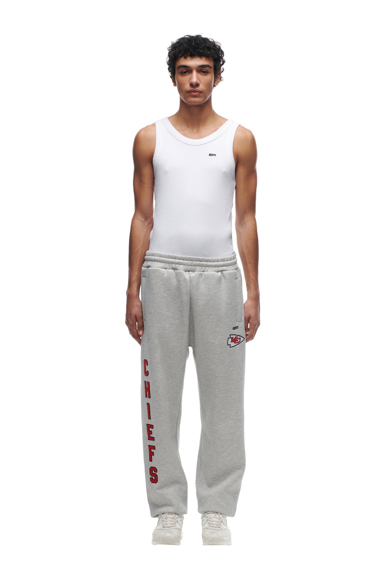 A man with curly hair wears the 6PM CHIEFS SEASON JOGGER, gray with CHIEFS and a sports logo on the left leg, paired with a white tank top and white sneakers, standing against a plain white background.