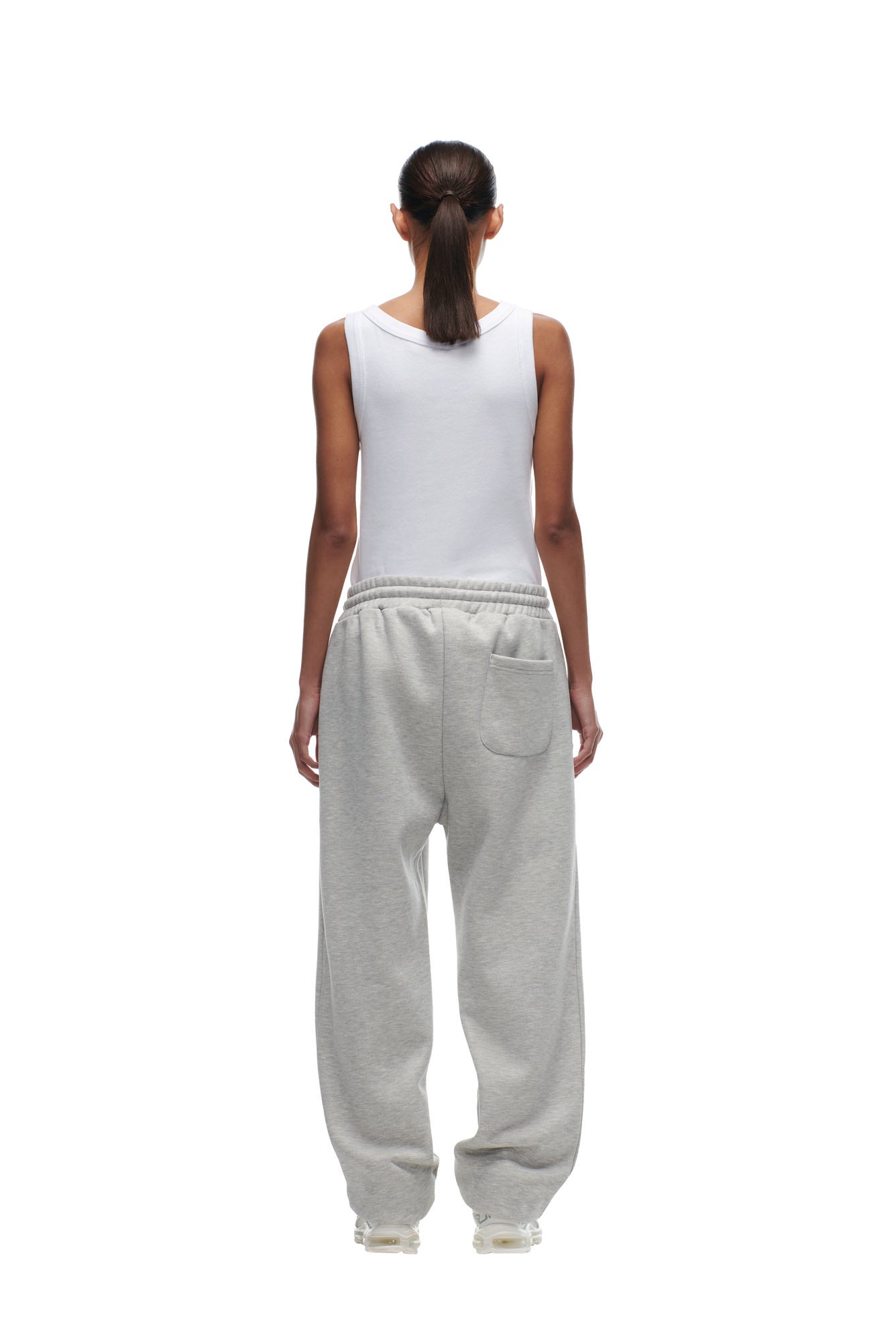 A woman with a ponytail faces away, wearing a white sleeveless top and loose, light gray CHIEFS SEASON JOGGER sweatpants by 6PM. The background is plain white.
