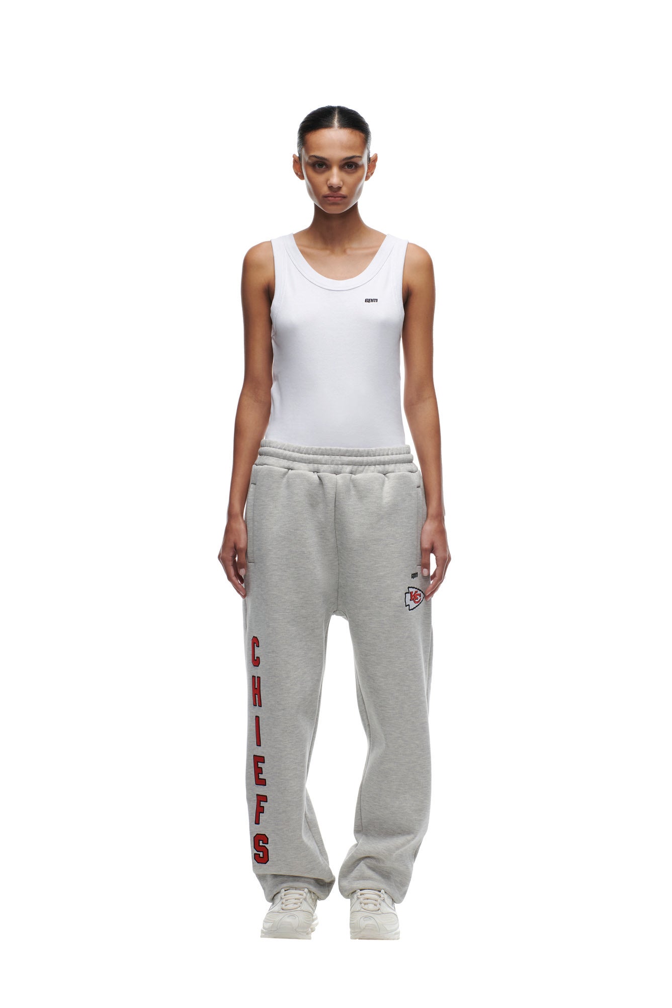 A woman faces forward in a white tank top and 6PM CHIEFS SEASON JOGGER gray sweatpants with red CHIEFS lettering and a small logo on the hip, paired with white sneakers, against a plain white background.