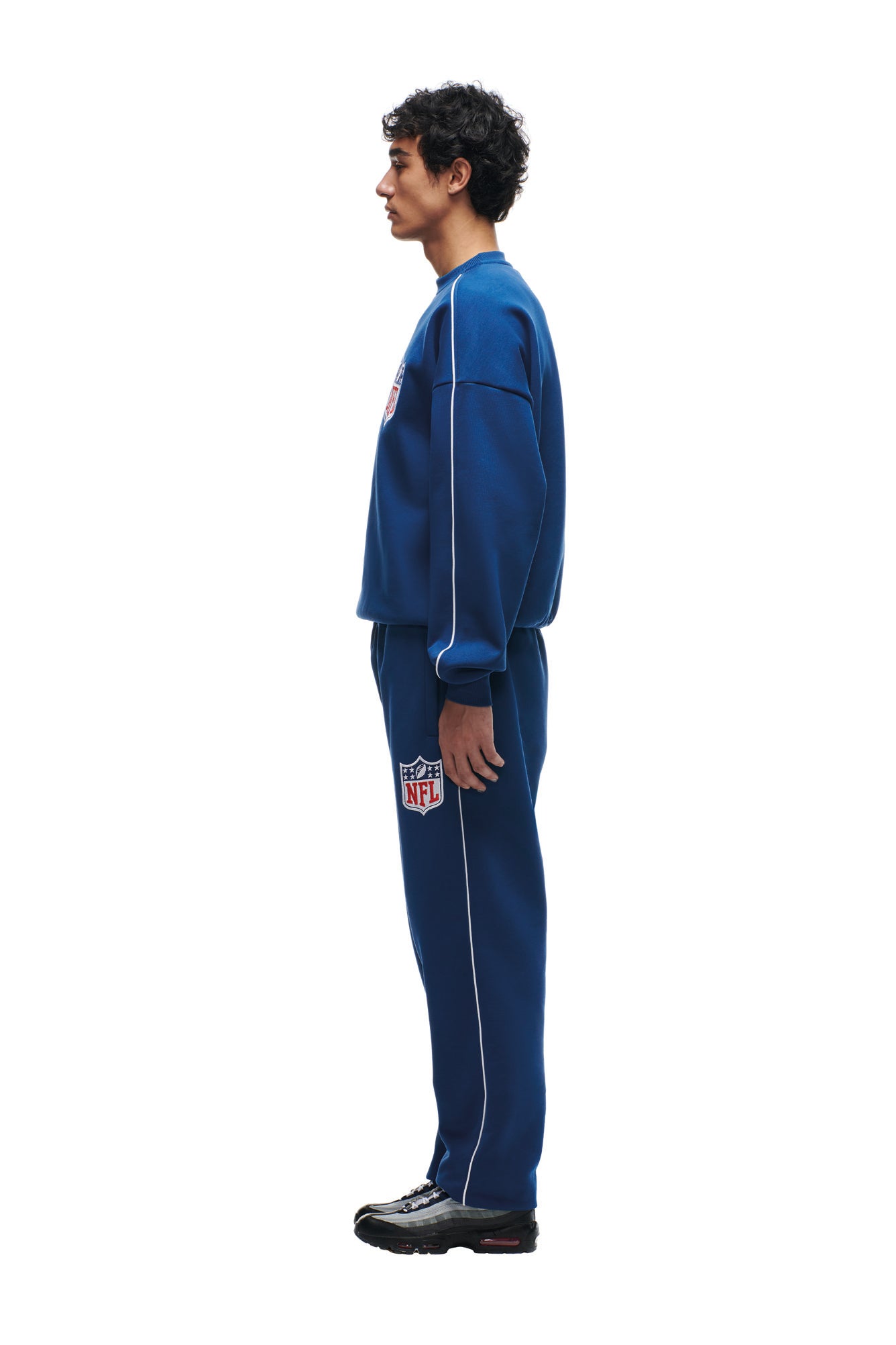 A person stands in profile wearing the 6PM SHIELD LOGO CREWNECK BLUE sweatshirt and matching sweatpants with NFL logos, black sneakers, and white side piping, against a plain white background.