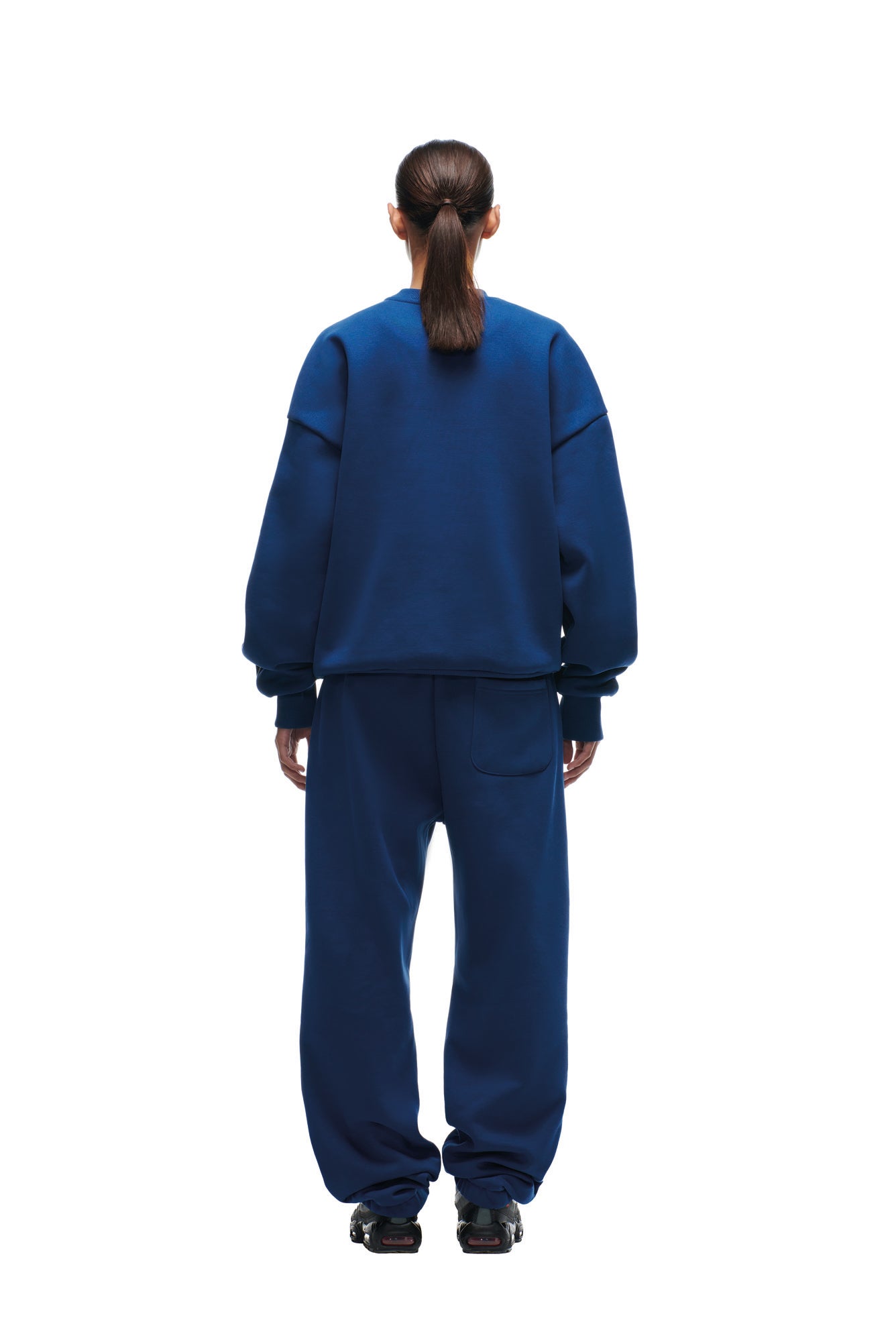 A person with long brown hair in a ponytail, wearing the 6PM SHIELD LOGO CREWNECK BLUE sweatshirt and matching sweatpants with black sneakers, stands facing away against a white background.