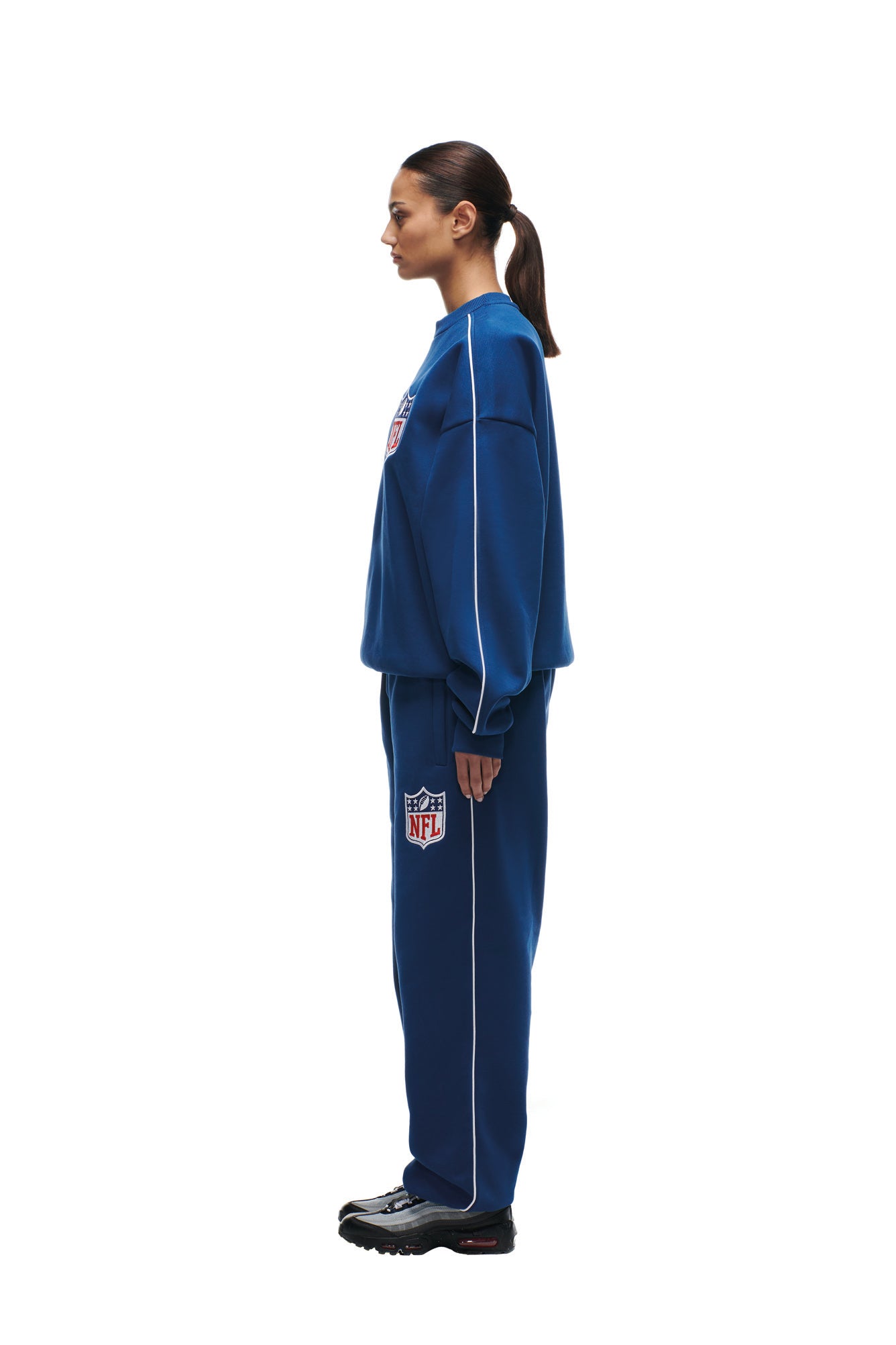 A woman stands in profile against a white background, wearing the 6PM SHIELD LOGO CREWNECK BLUE and black sneakers. Her hair is tied back in a low ponytail.