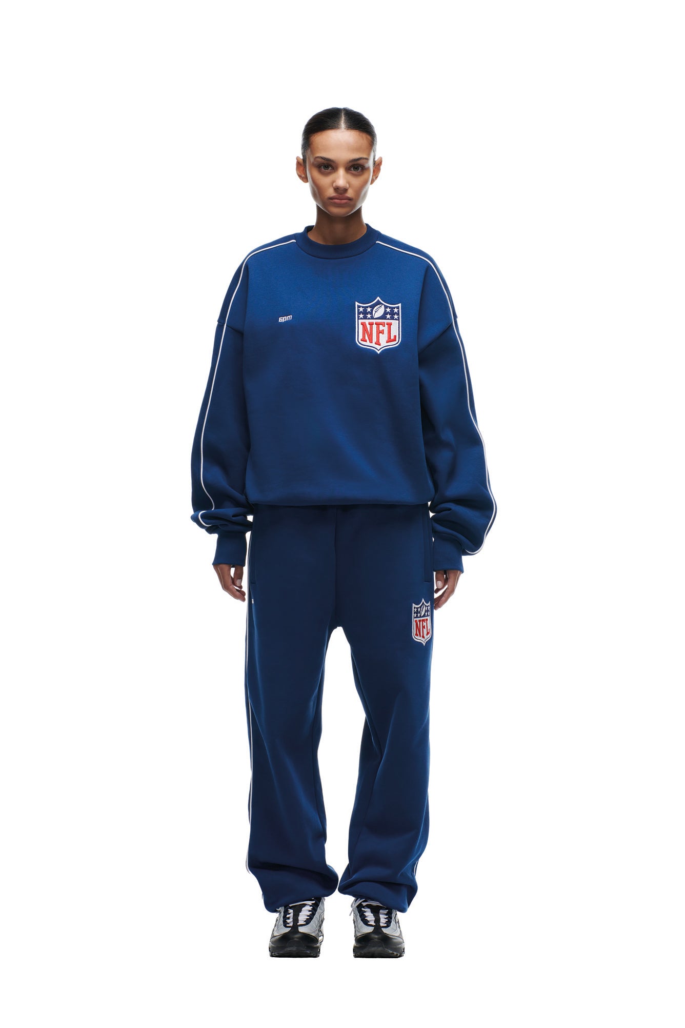 A person stands facing forward in a 6PM SHIELD LOGO CREWNECK BLUE set with white piping and NFL logos on both the sweatshirt and pants, hair slicked back, black sneakers, against a white background.