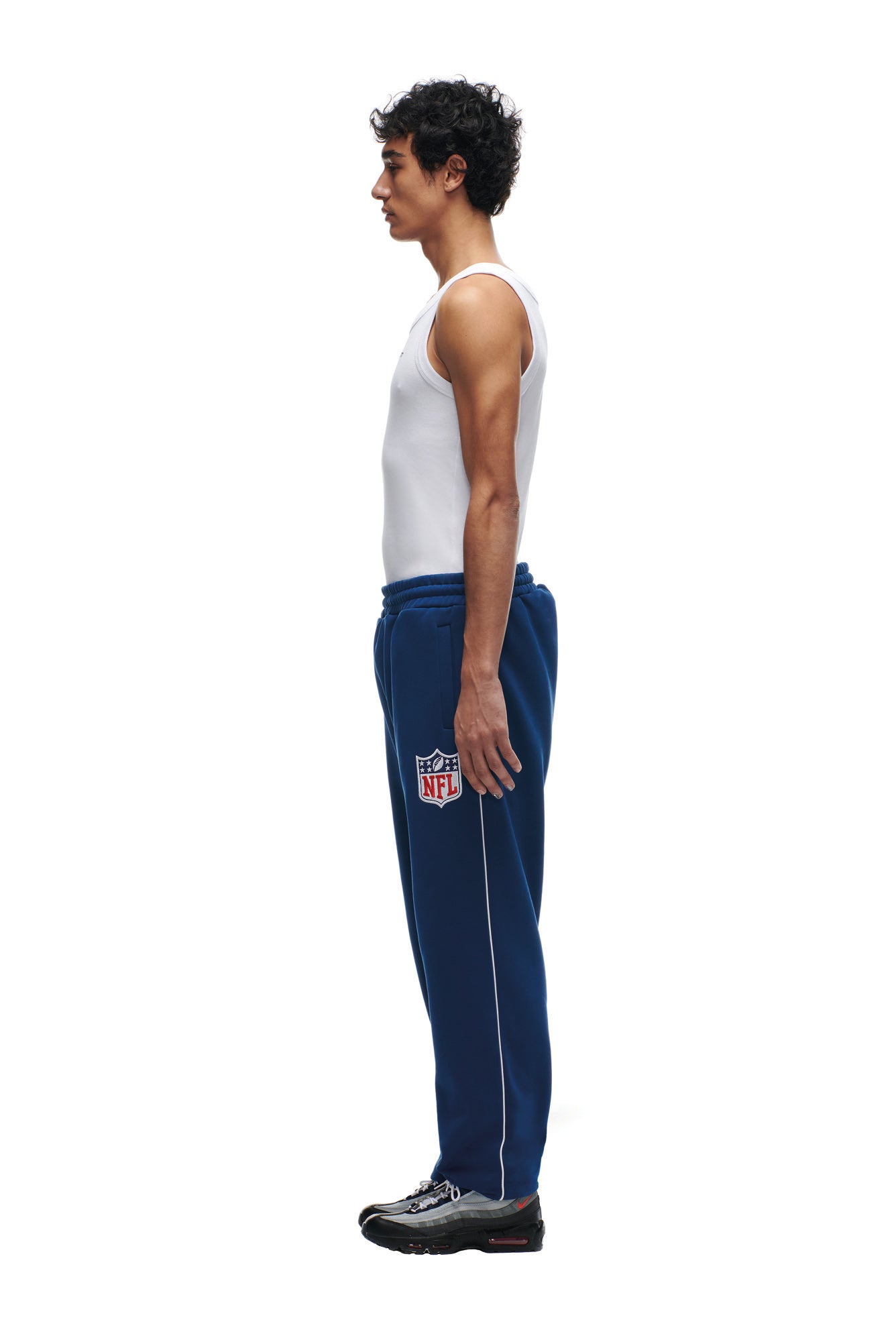 A man stands in profile against a white background, wearing a white tank top, 6PM SHIELD LOGO JOGGER BLUE sweatpants, and black sneakers.