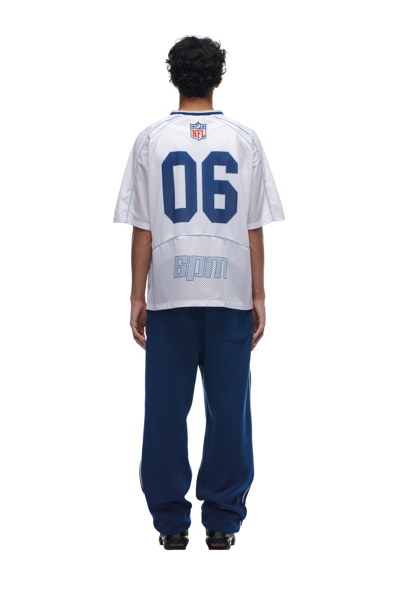 A person stands with their back to the camera, wearing the 6PM NFL MESH SHIRT in white, featuring "06" and "BPM" on the back, paired with loose blue pants and black shoes. The background is plain white.