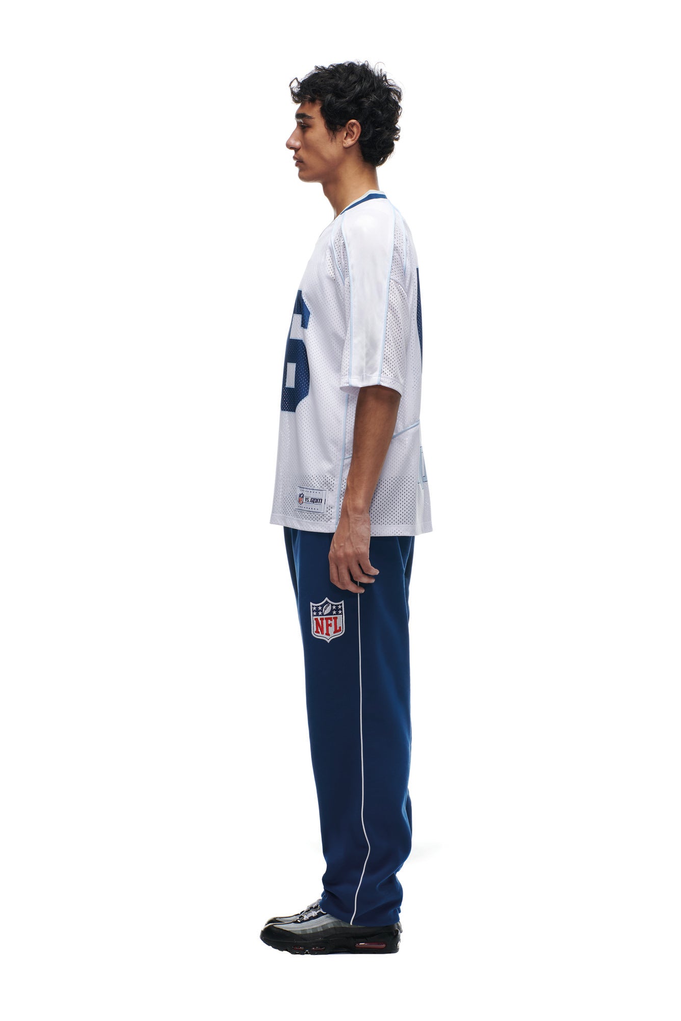 A young man stands in profile against a white background, wearing the 6PM NFL MESH SHIRT, blue NFL logo pants, and black athletic shoes.