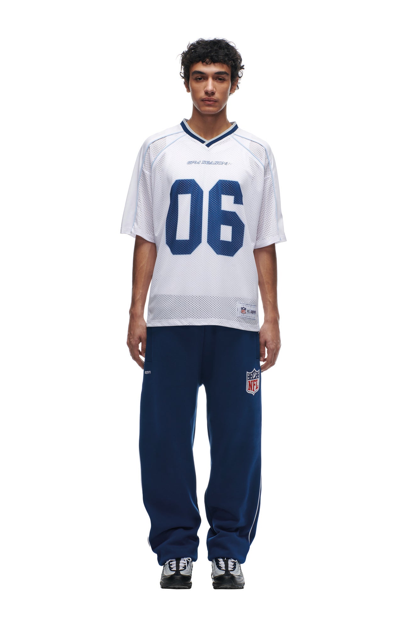 A person faces forward in a white 6PM NFL MESH SHIRT with "06" on the front, paired with navy blue NFL logo sweatpants and black sneakers. The background is plain white.