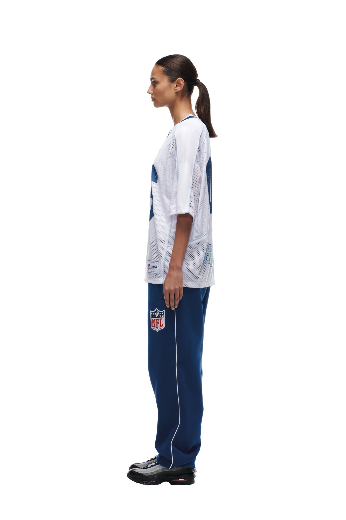 A woman in profile wears a 6PM NFL MESH SHIRT and blue NFL sweatpants with black sneakers. Her hair is tied back in a ponytail, and she stands against a plain white background.