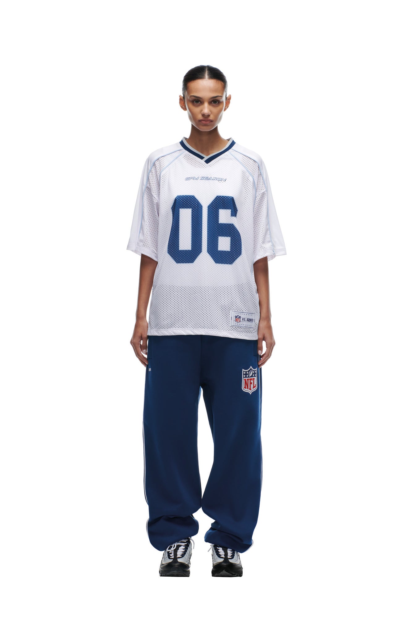 A woman faces forward in a loose white 6PM NFL MESH SHIRT with blue "06," styled with baggy blue NFL sweatpants and black sneakers, all set against a plain white background.