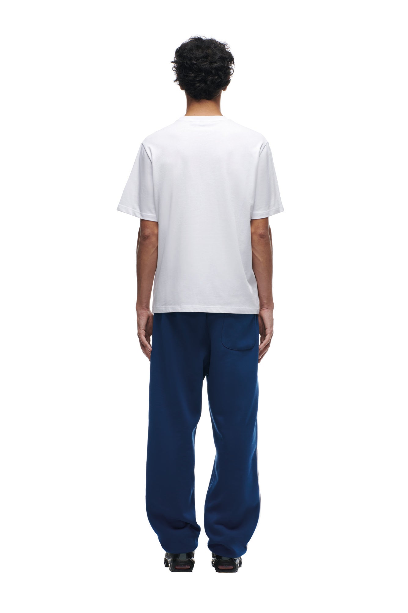 A person stands facing away from the camera against a white background, wearing a plain white NFL VS 6PM T-SHIRT by 6PM, loose blue pants, and black shoes.