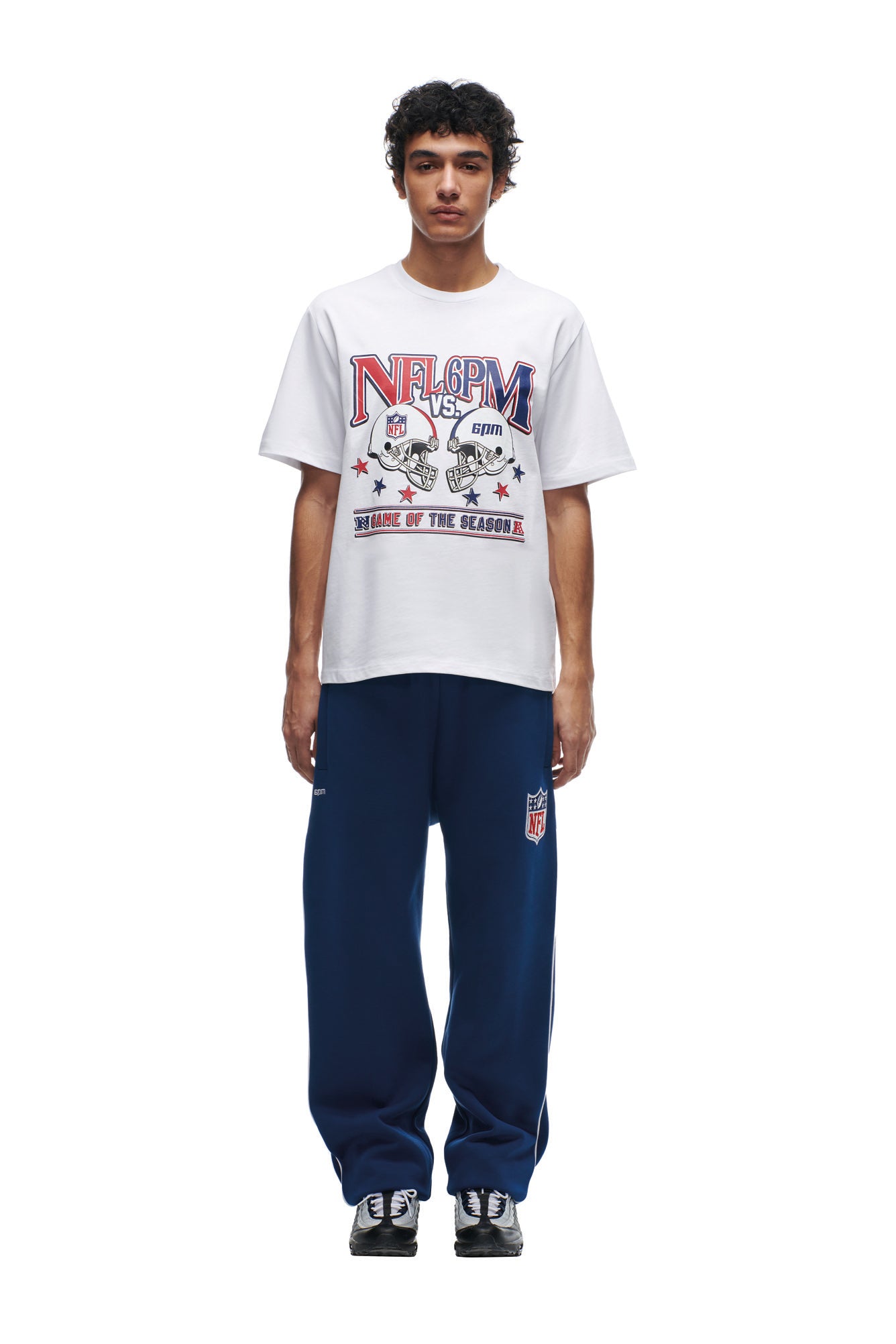 A person faces forward in a 6PM NFL VS 6PM T-SHIRT WHITE featuring a bold NFL graphic, paired with navy NFL sweatpants and black-and-white sneakers. The background is plain white.