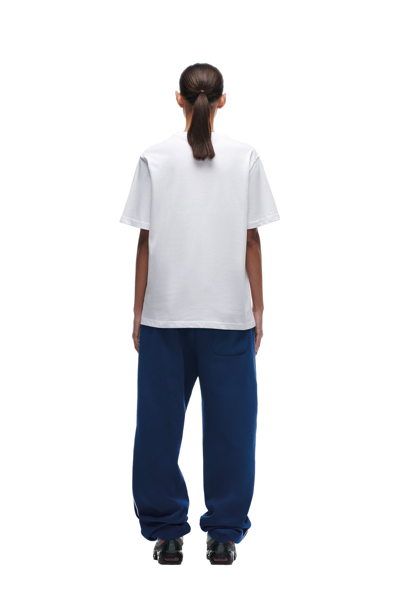 A person with long hair in a ponytail, wearing the 6PM NFL VS 6PM T-SHIRT WHITE and loose blue pants, stands facing away against a white background. Black shoes are visible.
