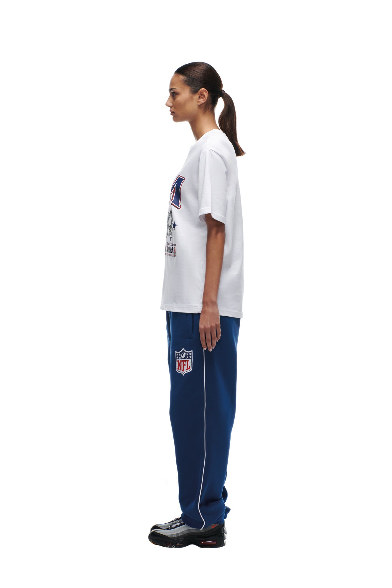 A woman stands in profile against a white background, wearing a 6PM NFL VS 6PM T-SHIRT in white, blue NFL sweatpants with logo, and black sneakers. Her hair is tied back in a low ponytail.