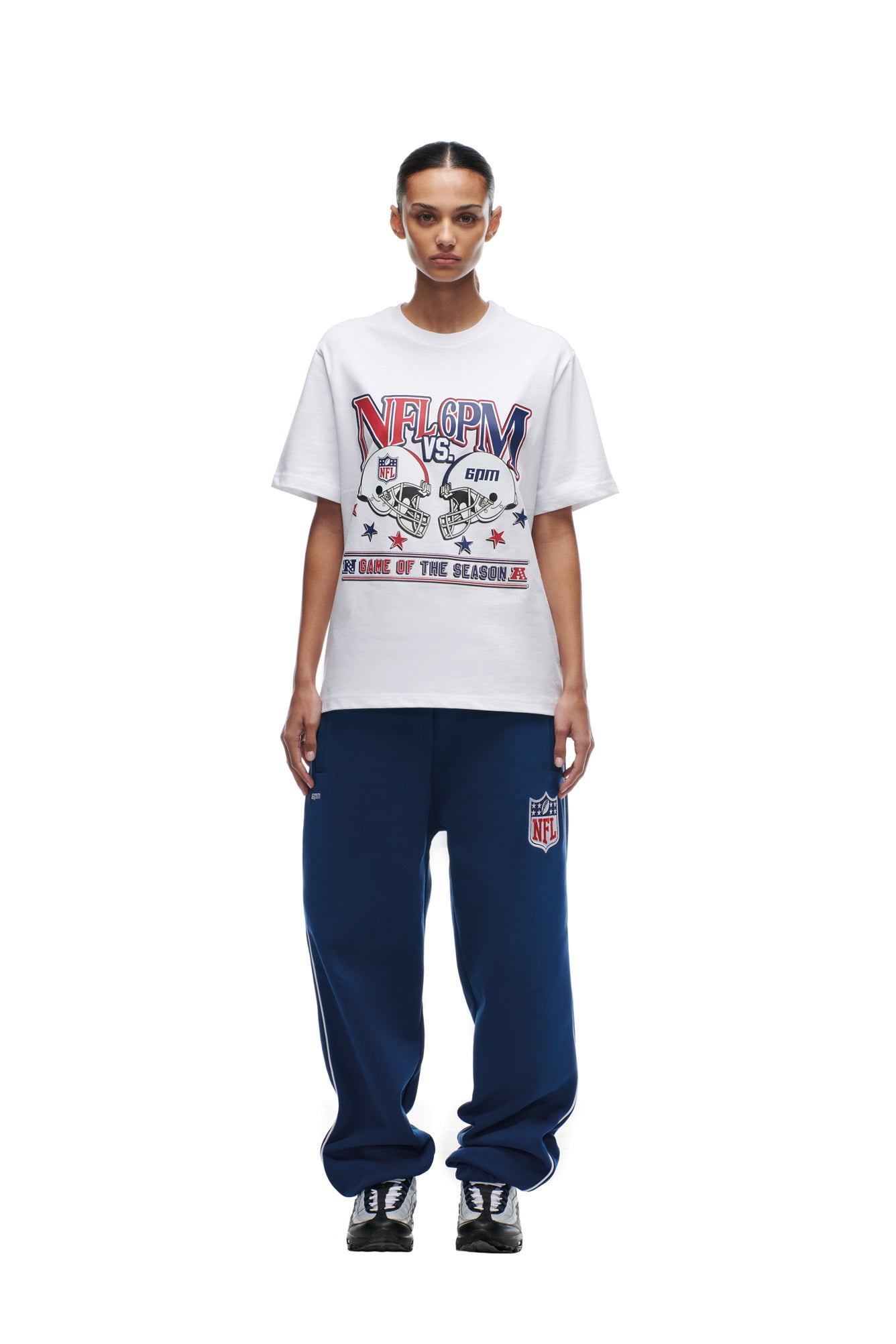 A person faces forward wearing a white 6PM NFL VS 6PM T-SHIRT and loose blue NFL sweatpants with black sneakers, standing against a plain white background.