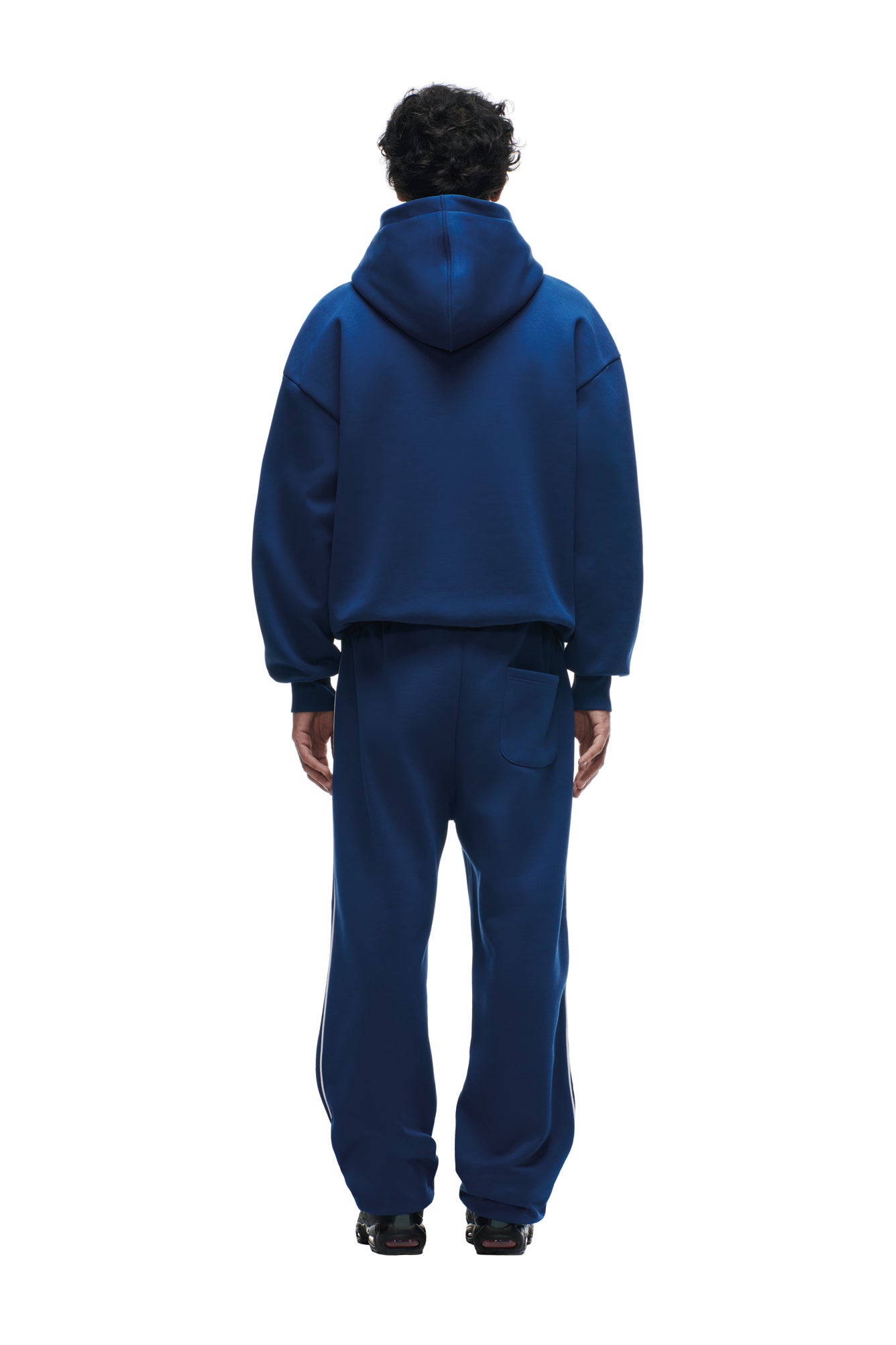 A person with short, dark curly hair faces away, wearing the 6PM SHIELD LOGO HOODIE BLUE and matching sweatpants against a plain white background.