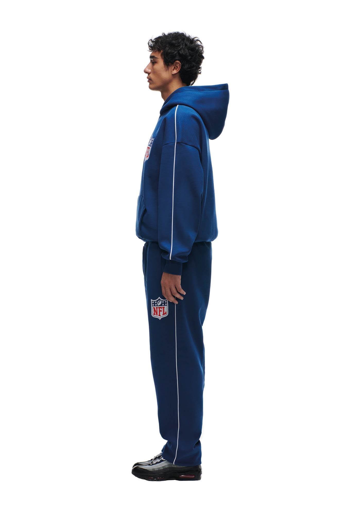 A person in profile wears the 6PM SHIELD LOGO HOODIE BLUE and matching blue sweatpants with NFL and REJECTS patches, paired with black sneakers, against a white background.