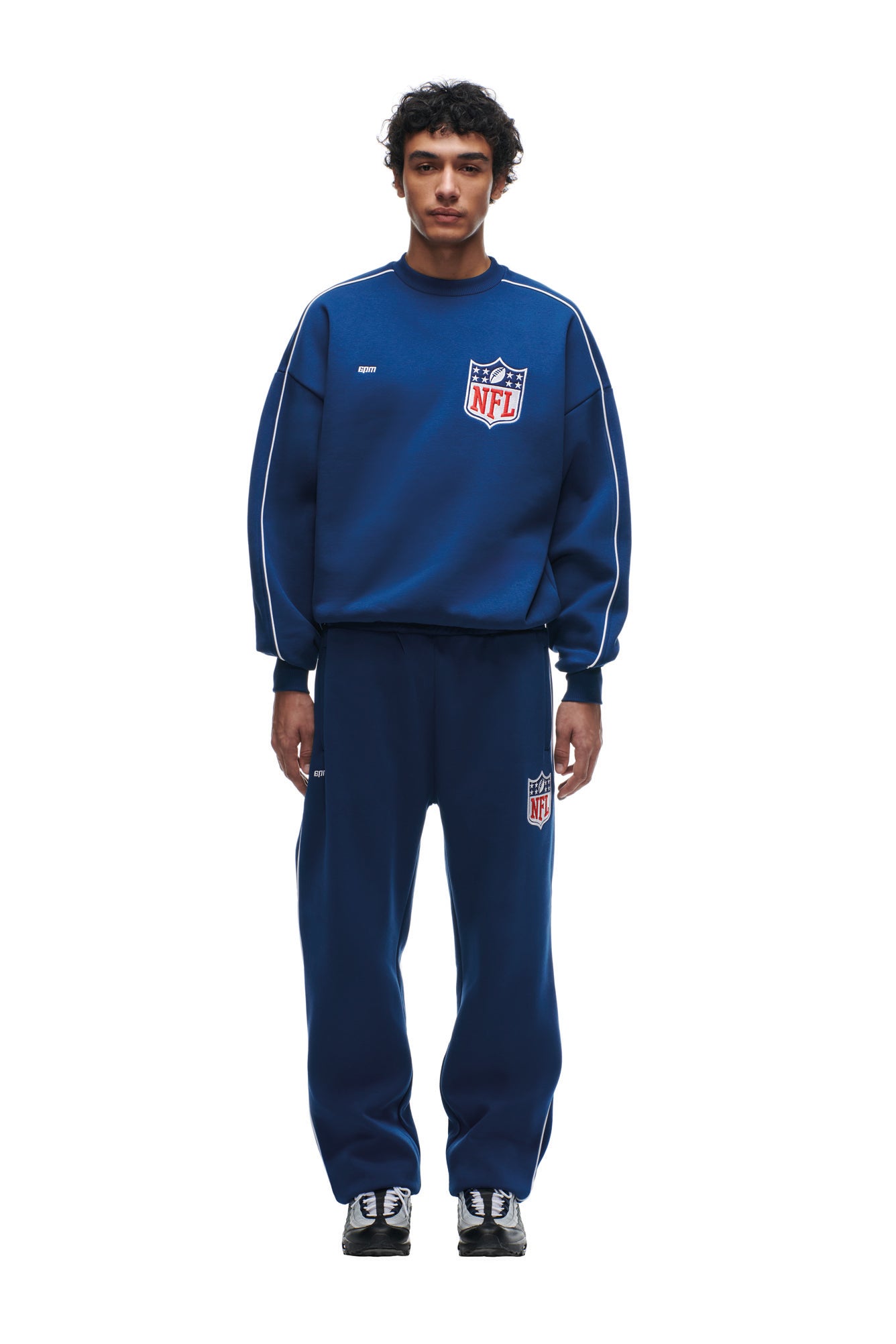 A person faces forward in a loose-fitting 6PM SHIELD LOGO CREWNECK BLUE sweatshirt and matching sweatpants, paired with black and white sneakers against a plain white background.