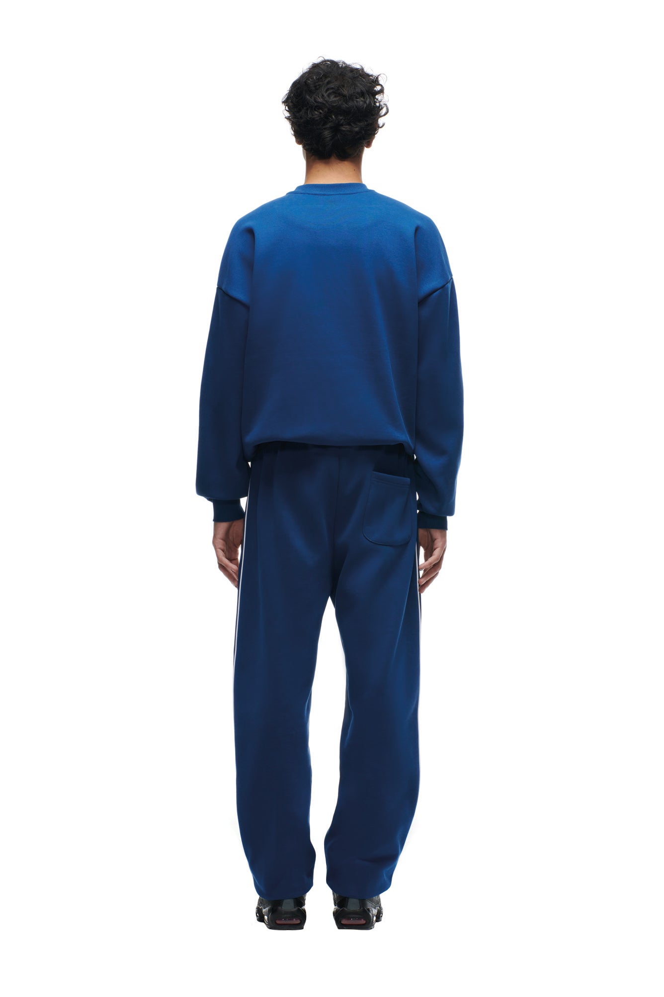 A person with short curly hair faces away, wearing the 6PM SHIELD LOGO CREWNECK BLUE sweatshirt and matching pants with black shoes, set against a plain white background.