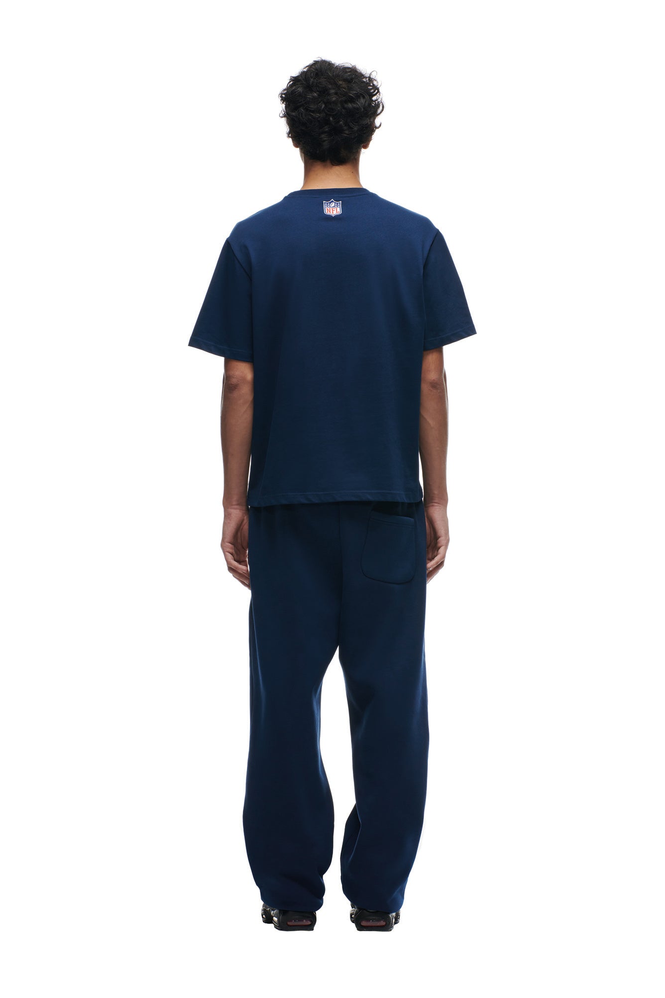 A person with short curly hair faces away, wearing a loose-fitting 6PM PATRIOTS SEASON T-SHIRT and matching navy blue pants, set against a plain white background.