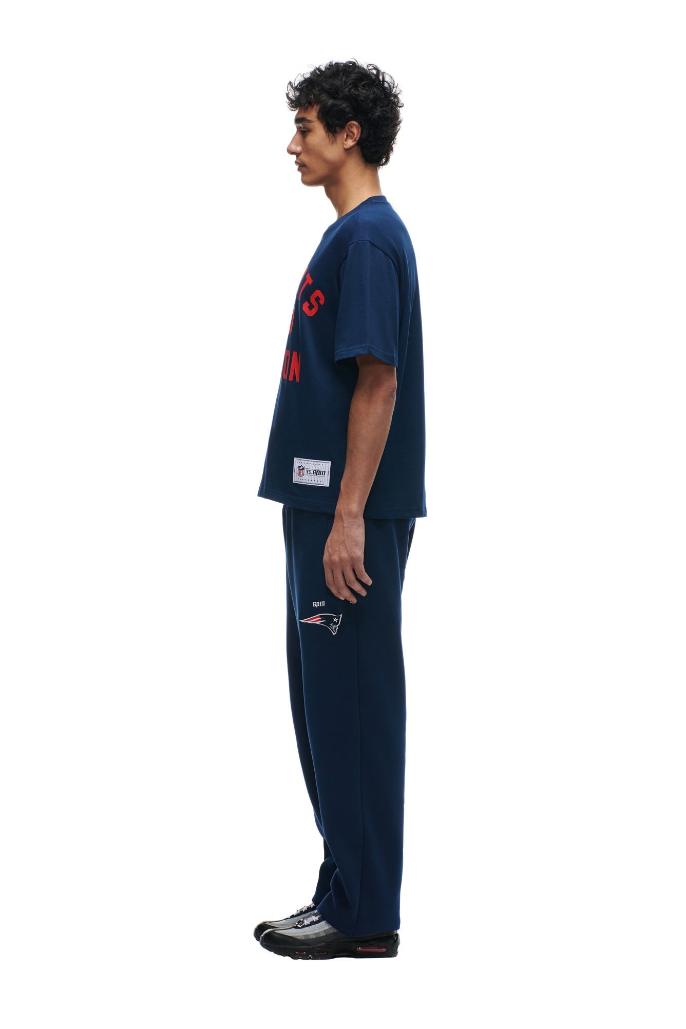 A man in profile wears the 6PM PATRIOTS SEASON T-SHIRT in navy with red text, paired with matching navy track pants featuring a small sports logo and black sneakers, set against a plain white background.