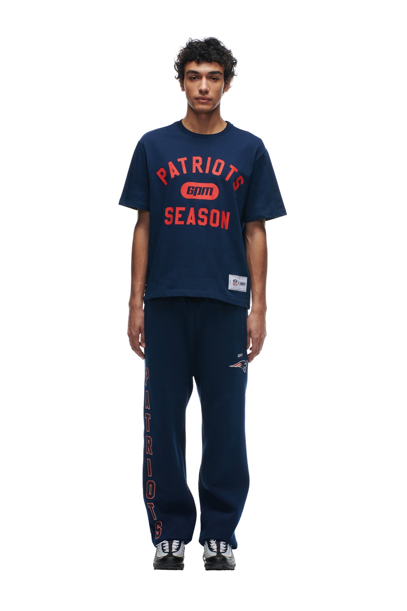 A person stands facing forward in a 6PM PATRIOTS SEASON T-SHIRT, navy sweatpants with "Patriots" vertically on one leg, and black and white sneakers.