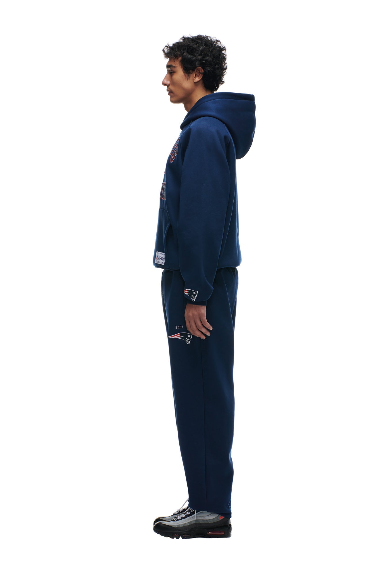 A man in profile models the 6PM PATRIOTS SEASON HOODIE and matching sweatpants, featuring NFL and New England Patriots logos, with black sneakers, set against a white background.
