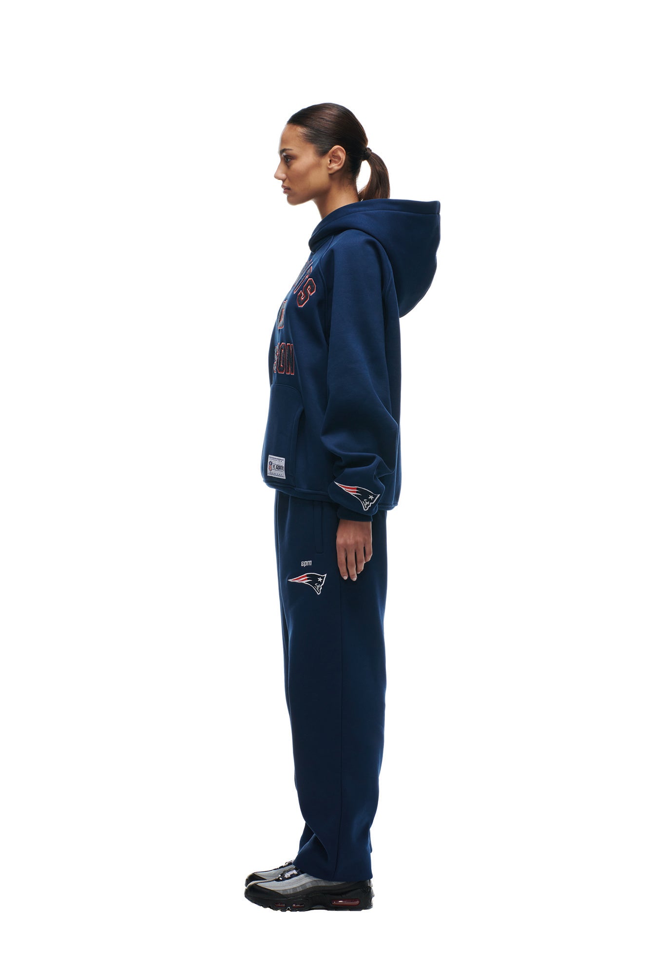 A woman in profile wears the 6PM PATRIOTS SEASON HOODIE and matching sweatpants with sports team logos, paired with black sneakers, against a plain white background.