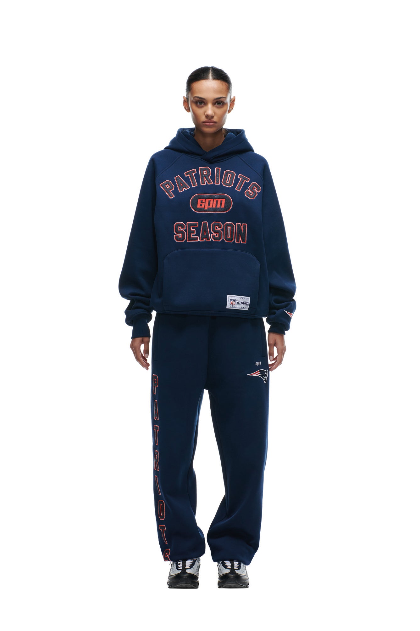 A person faces forward in a PATRIOTS SEASON HOODIE by 6PM, navy sweatpants with "Patriots" down one leg, and black sneakers, all set against a white background.