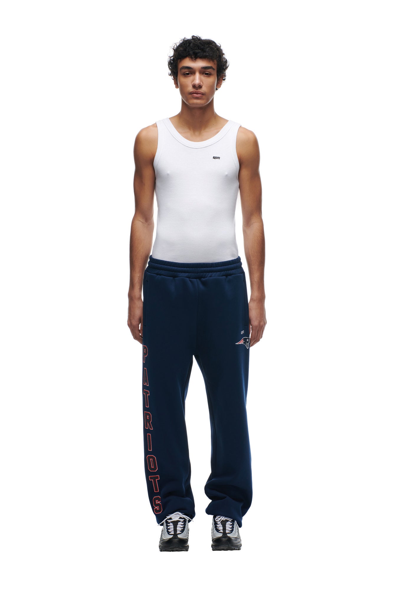 A person stands facing forward in a white tank top, 6PM PATRIOTS SEASON JOGGER navy sweatpants with "PATRIOTS" down one leg, and black-and-white sneakers against a plain white background.