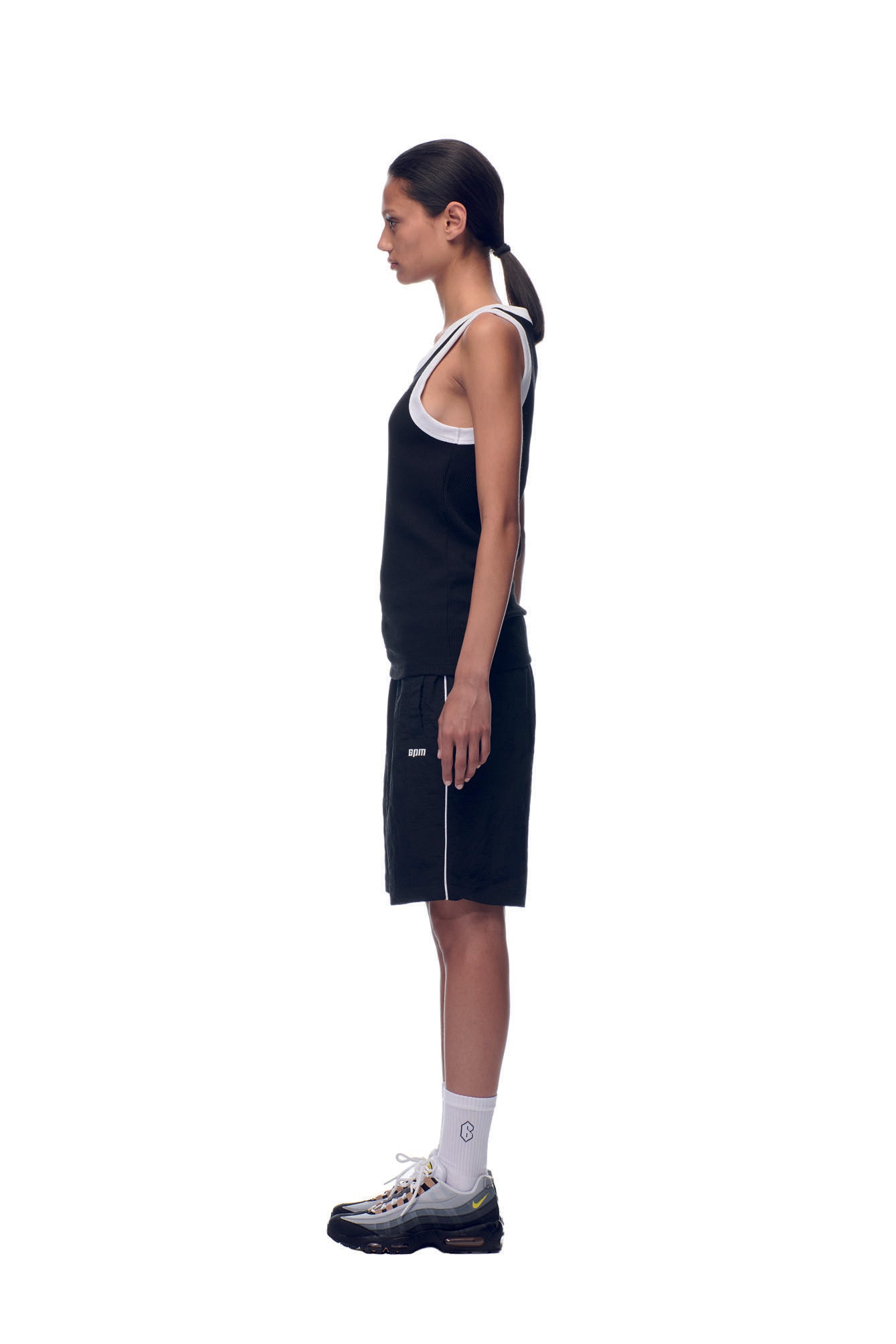 A person in profile stands against a white background, wearing the 6PM NH TANKTOP BLACK/WHITE, black shorts, white socks, black sneakers, and has a low ponytail.