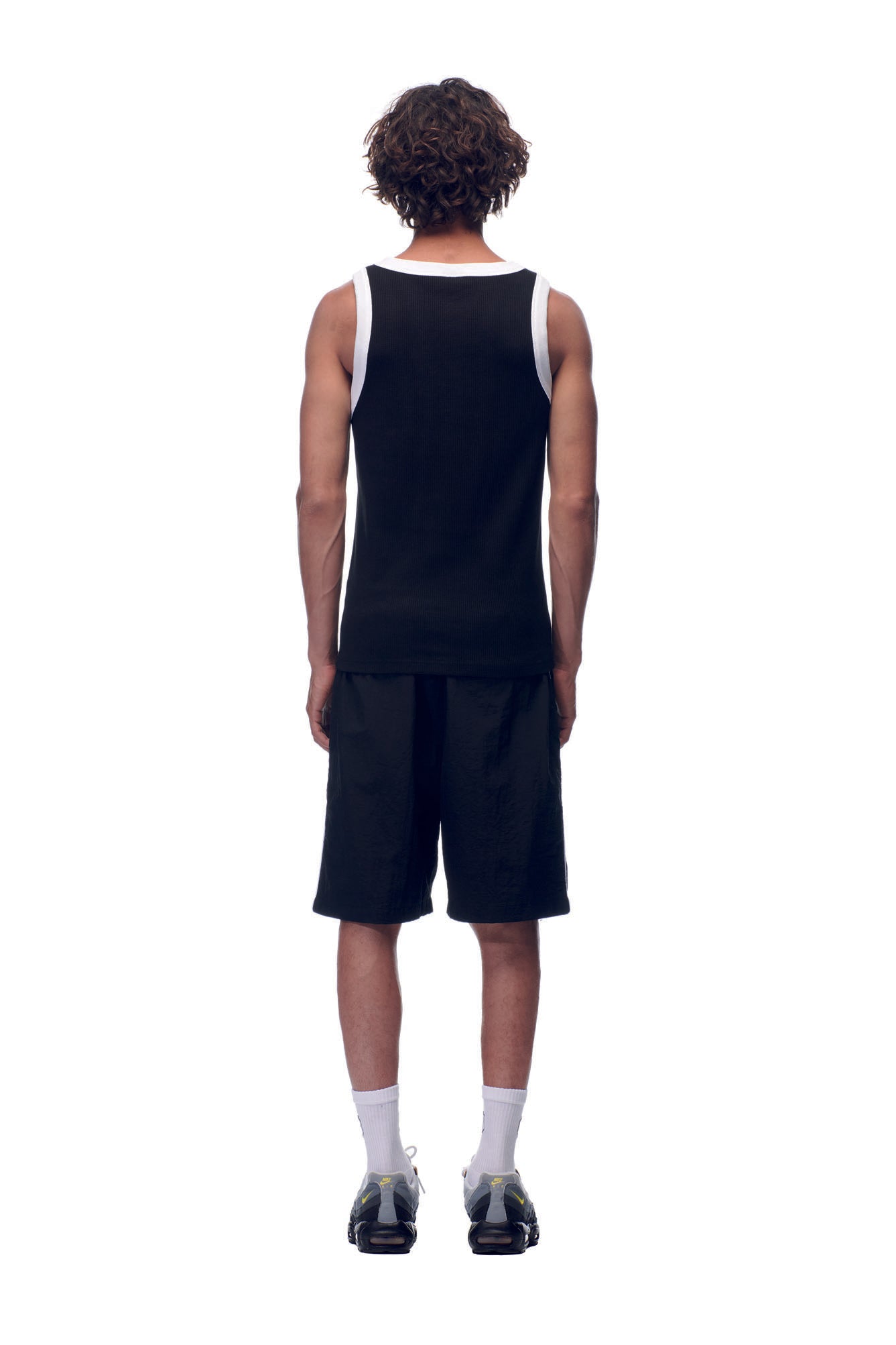 A person with curly hair, seen from behind, wears the 6PM NH TANKTOP BLACK/WHITE along with black shorts, white socks, and black sneakers, standing against a plain white background.