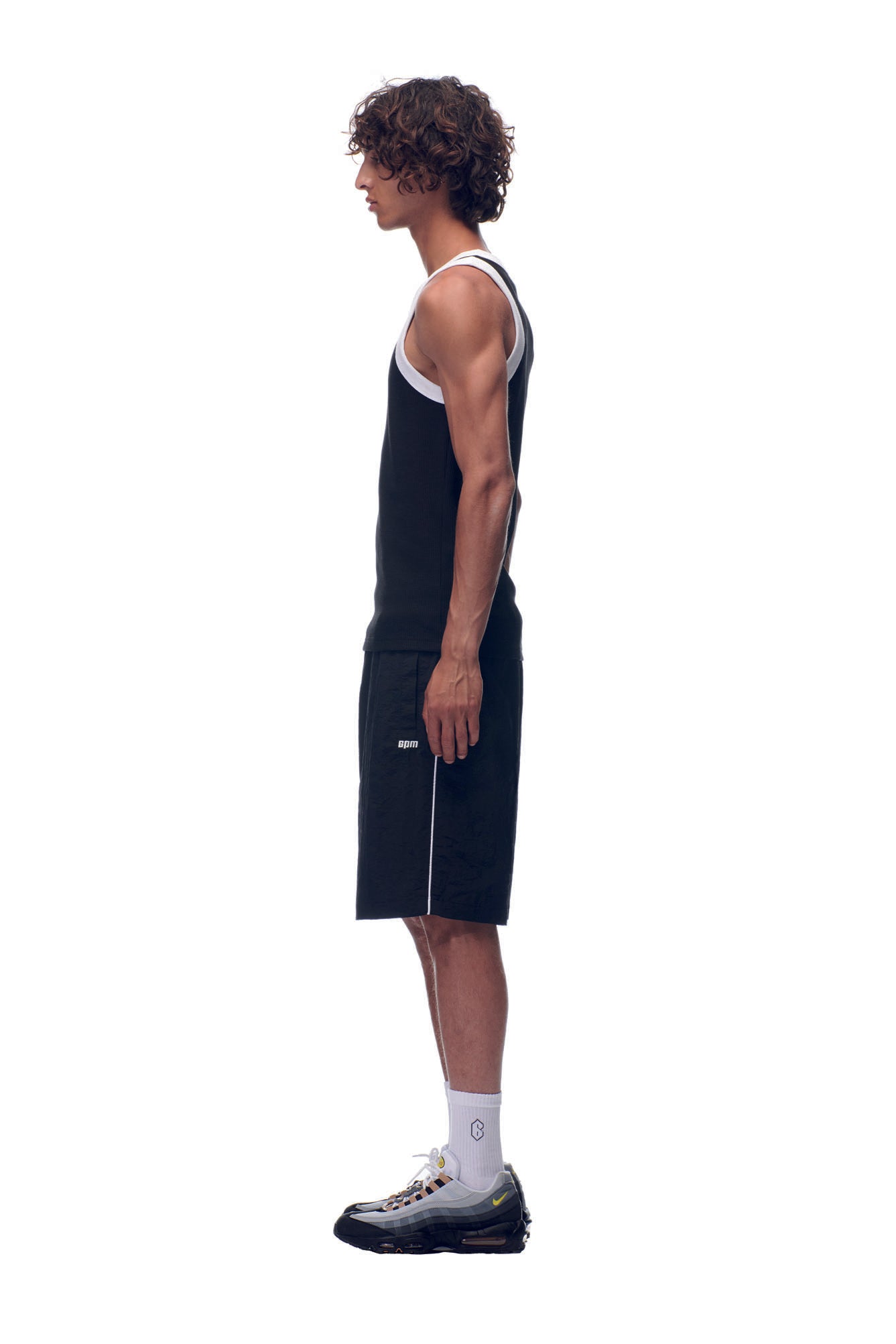 A young man with curly hair stands in profile against a white background, wearing the 6PM NH TANKTOP BLACK/WHITE, along with black shorts with white trim, white socks, and black sneakers.