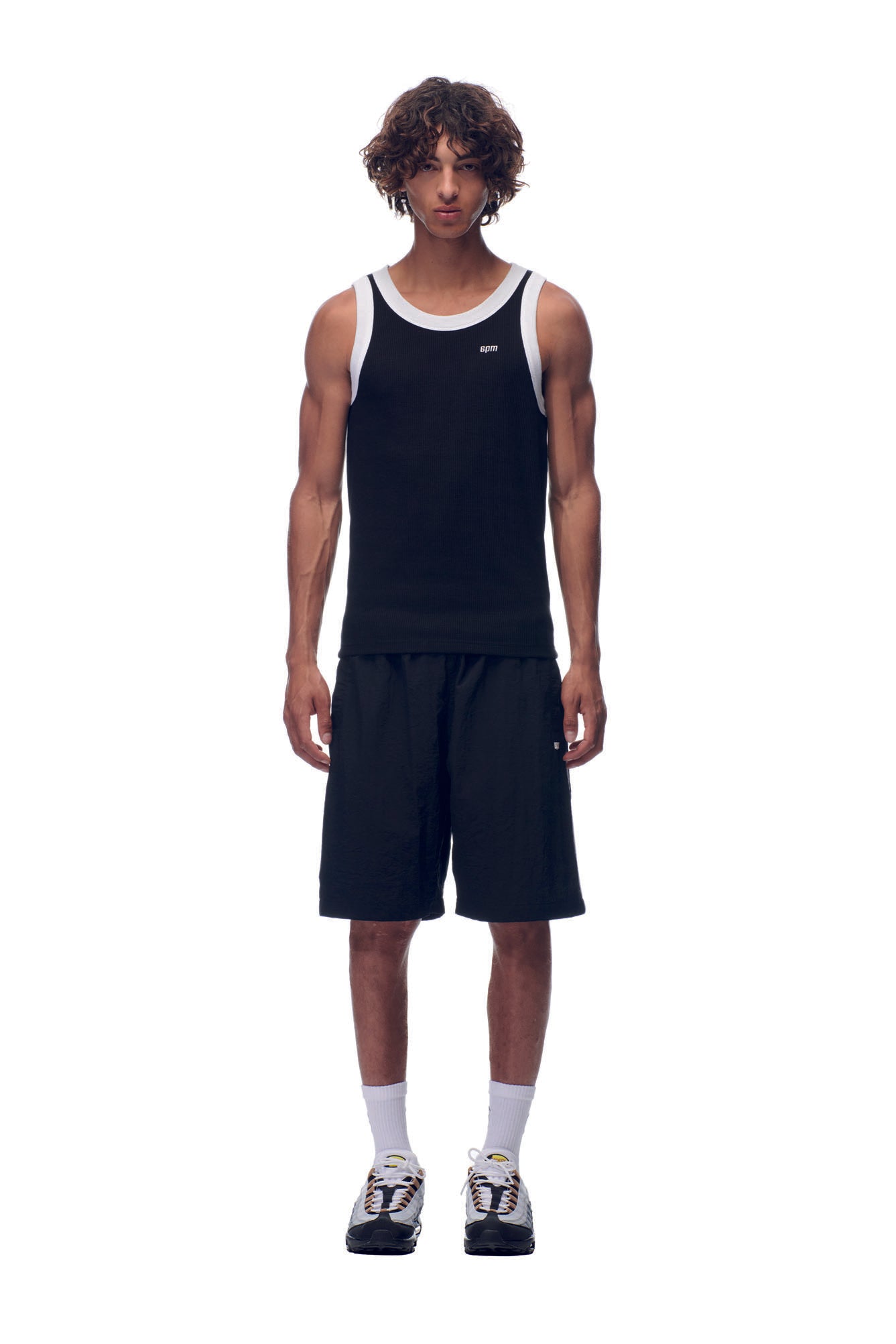 A man with curly hair faces forward, wearing the 6PM NH TANKTOP BLACK/WHITE, black shorts, white socks, and black and white sneakers, all set against a plain white background.