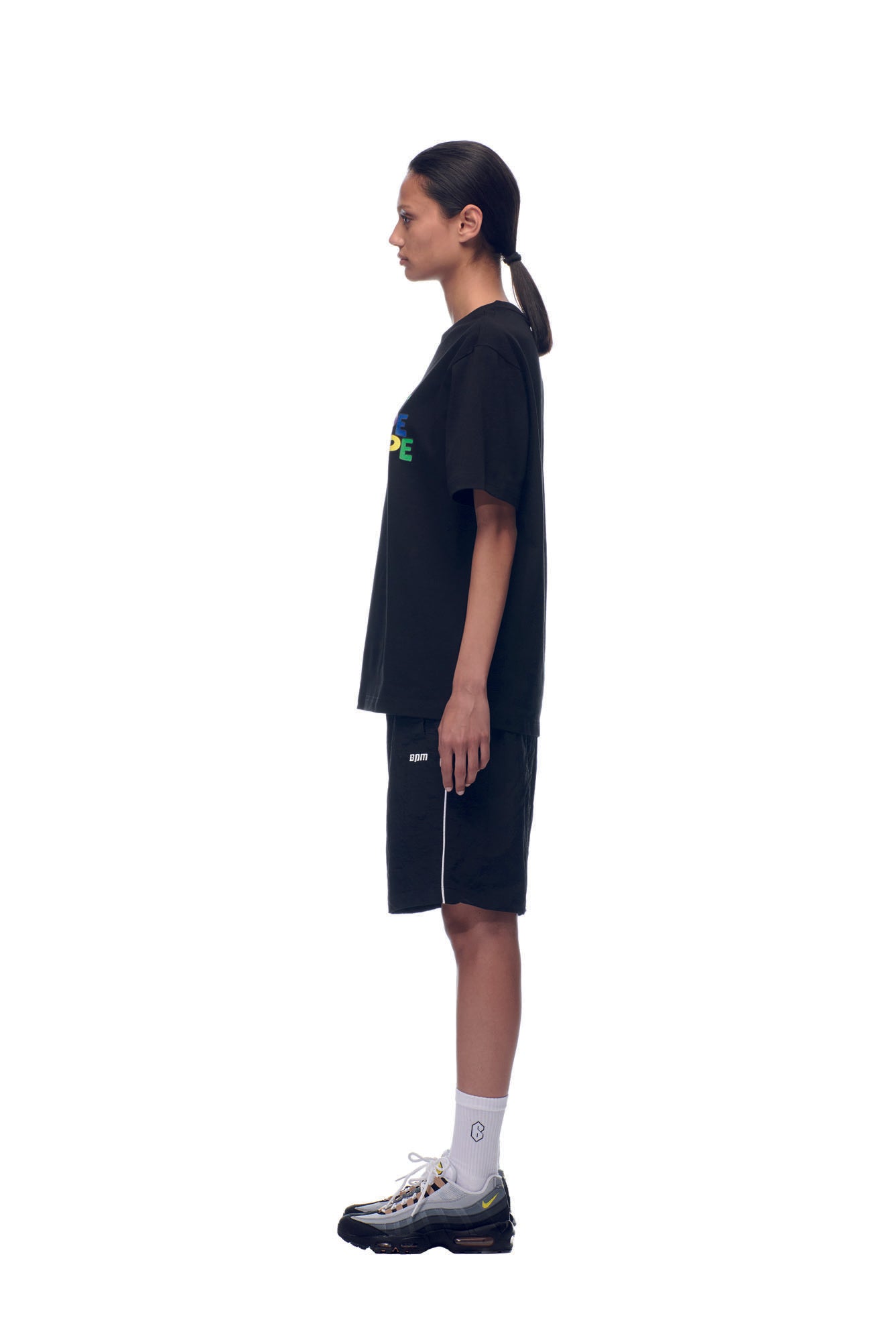A person stands in profile against a white background, wearing the 6PM SELL HOPE T-SHIRT BLACK along with black shorts, white crew socks, and black sneakers. Their hair is tied back in a low ponytail.