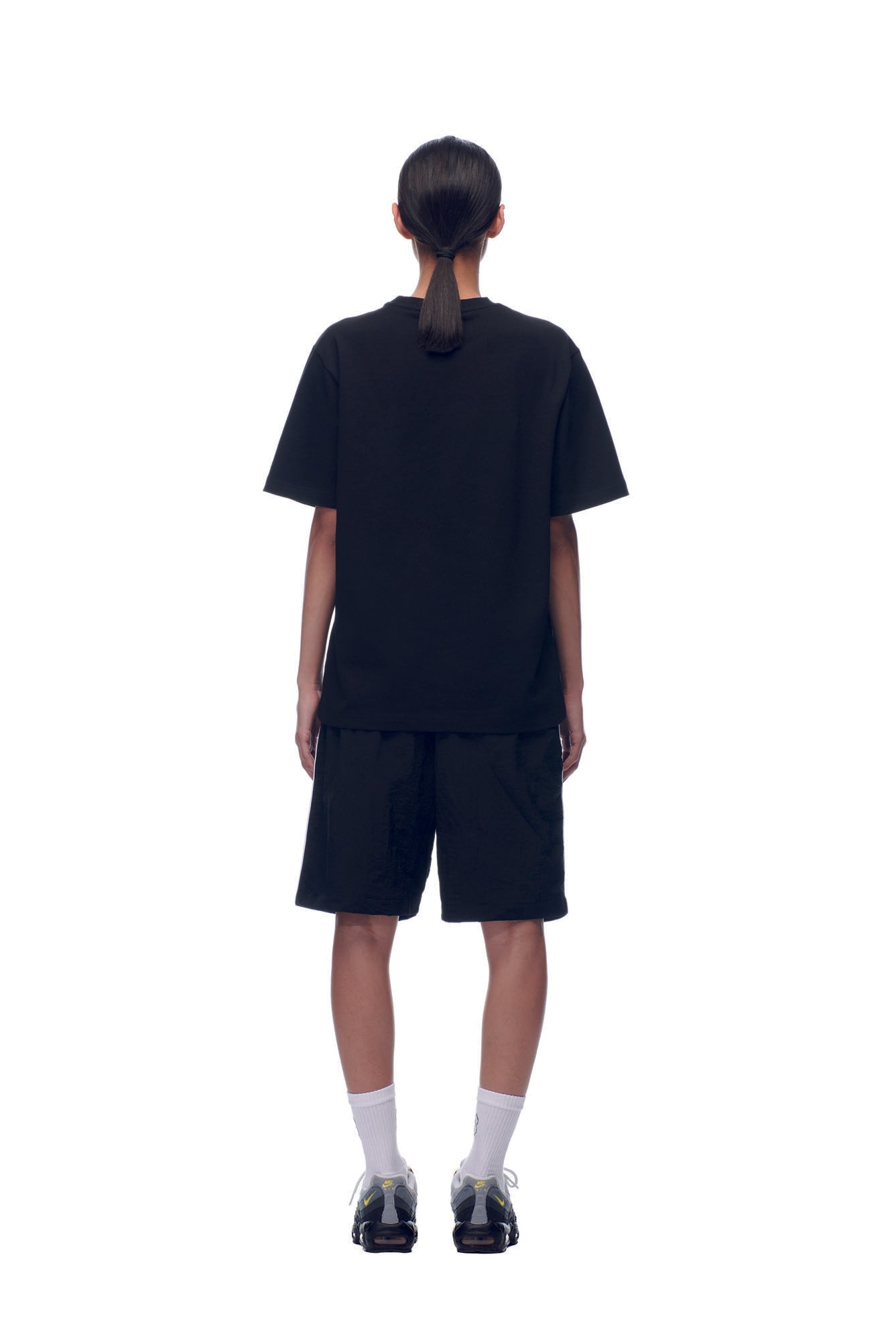A person with hair in a low ponytail stands facing away from the camera against a white background, wearing the 6PM NH PLAY LOGO T-SHIRT BLACK, black shorts, white socks, and black sneakers.
