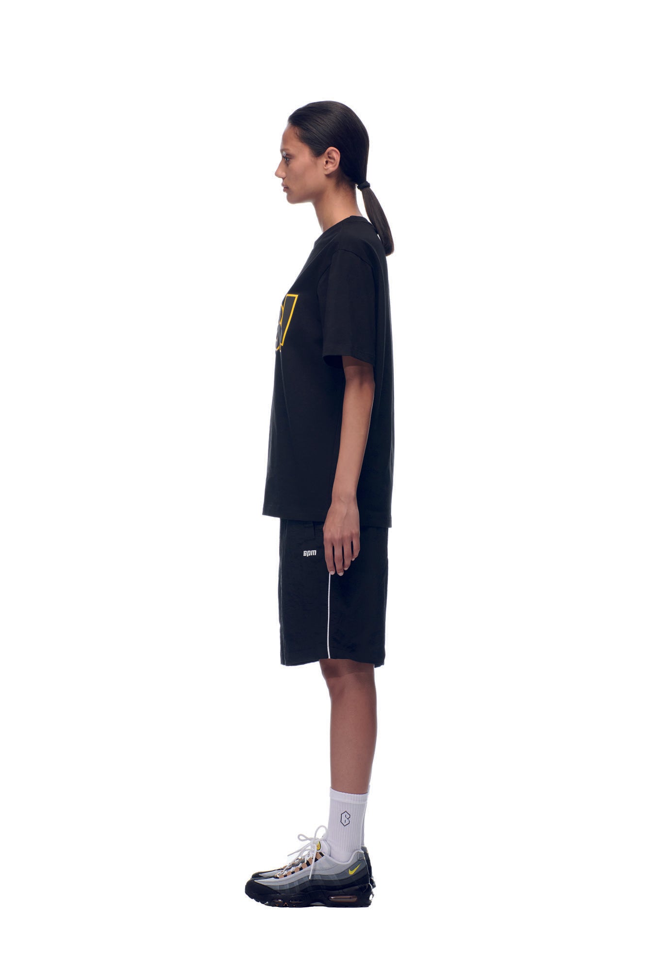 A person with straight dark hair in a low ponytail wears a 6PM NH PLAY LOGO T-SHIRT BLACK, black shorts, white socks, and black sneakers, standing in profile against a white background.