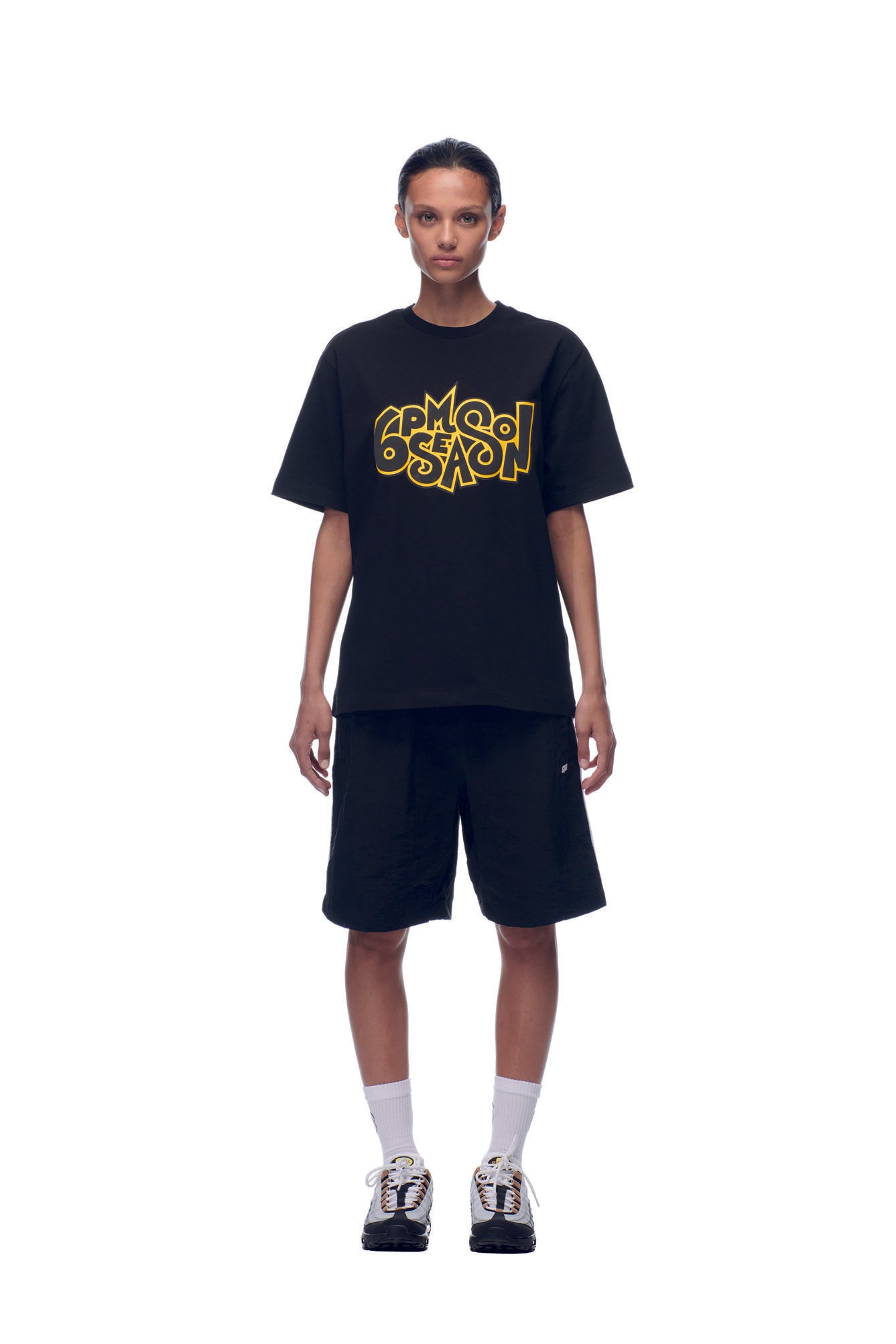 A person stands facing forward against a plain white background, wearing the 6PM NH PLAY LOGO T-SHIRT BLACK, black knee-length shorts, white socks, and black-and-white sneakers.