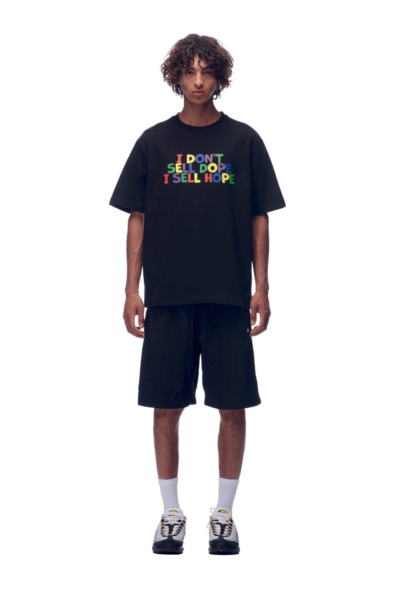A person faces forward against a white background, wearing the 6PM SELL HOPE T-SHIRT BLACK with colorful "I DON’T SELL DOPE I SELL HOPE" text, paired with black shorts, white socks, and sneakers.