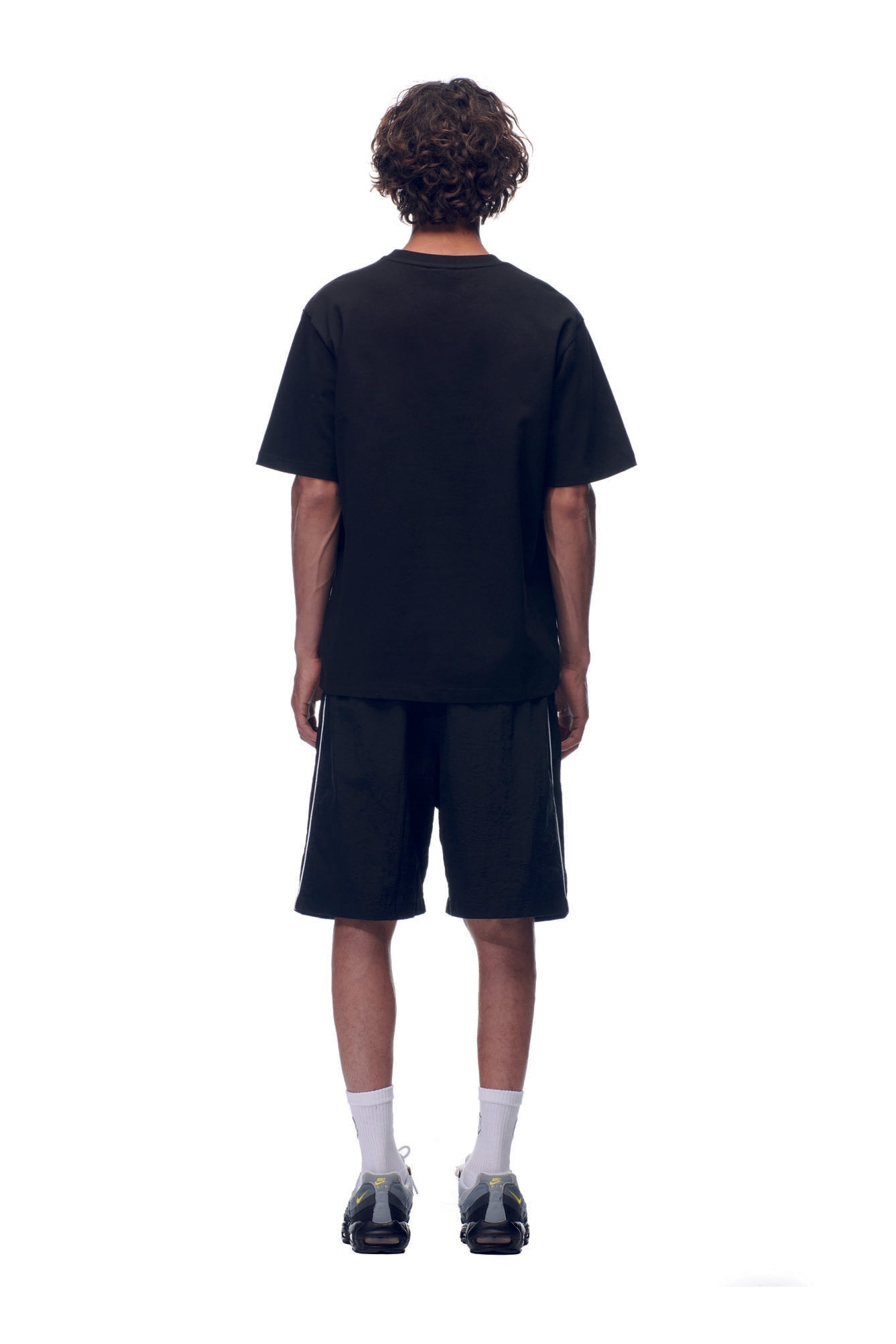 A person with curly hair, seen from behind, wears the 6PM NH PLAY LOGO T-SHIRT BLACK with black shorts, white socks, and black sneakers against a plain white background.