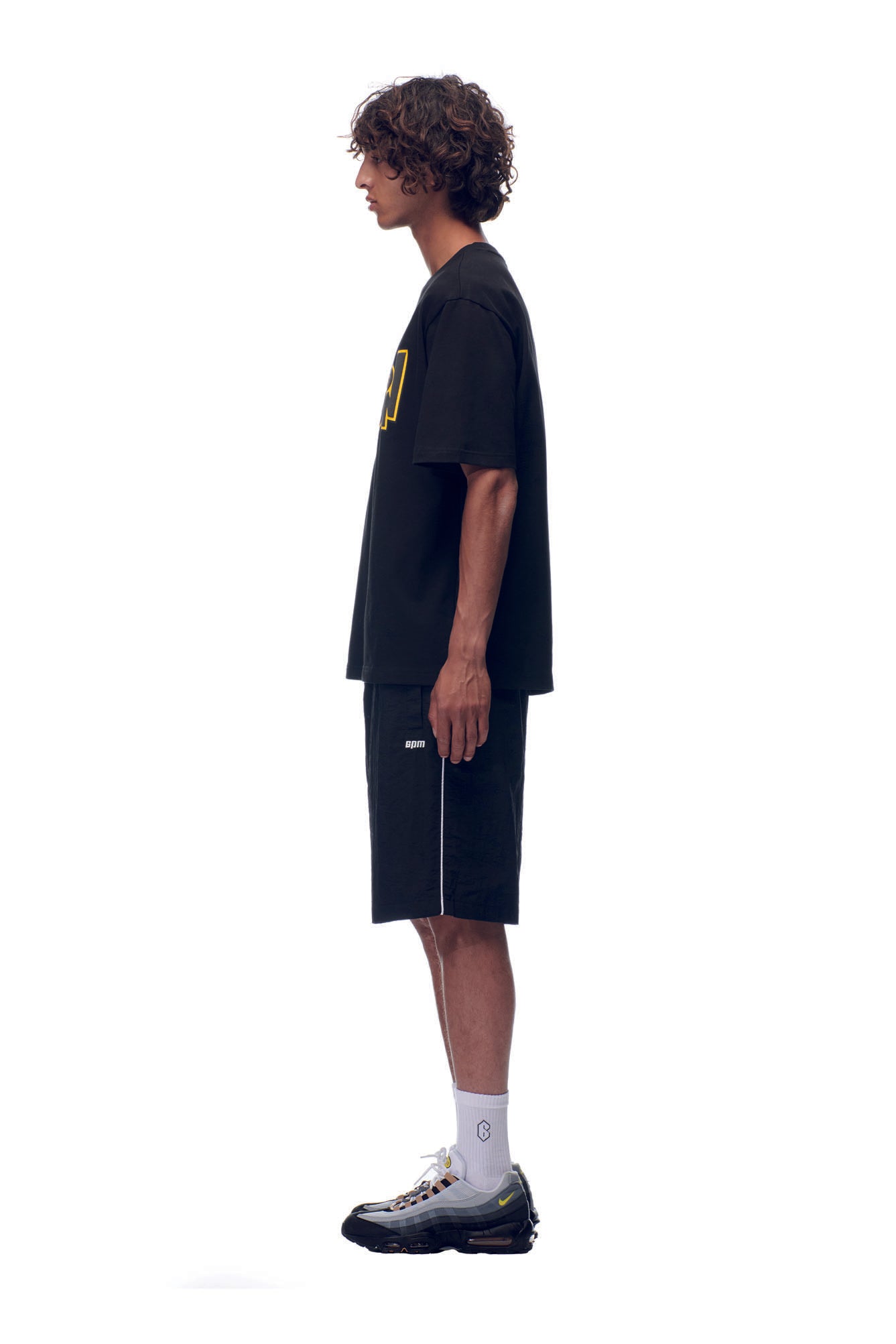 A man with curly hair stands in profile against a white background, wearing the 6PM NH PLAY LOGO T-SHIRT BLACK, black shorts, white socks, and black sneakers with yellow accents.