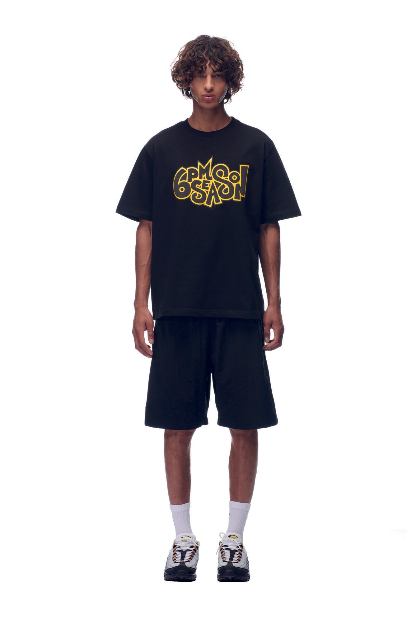 A young man with curly hair wears the 6PM NH PLAY LOGO T-SHIRT BLACK, paired with black shorts, white socks, and sneakers, standing against a plain white background.