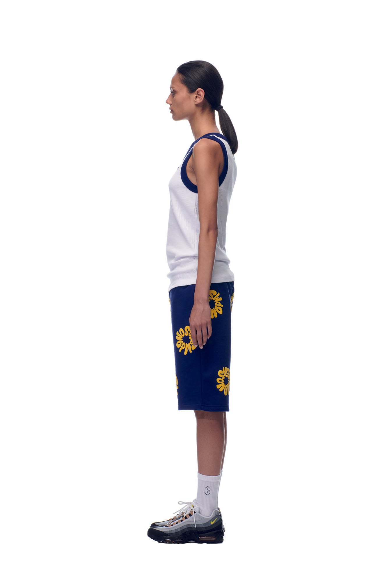 A person stands in profile against a white background, wearing the 6PM NH Tanktop White/Navy, blue shorts with yellow sun designs, white socks, and black athletic shoes. Their hair is tied back in a low ponytail.