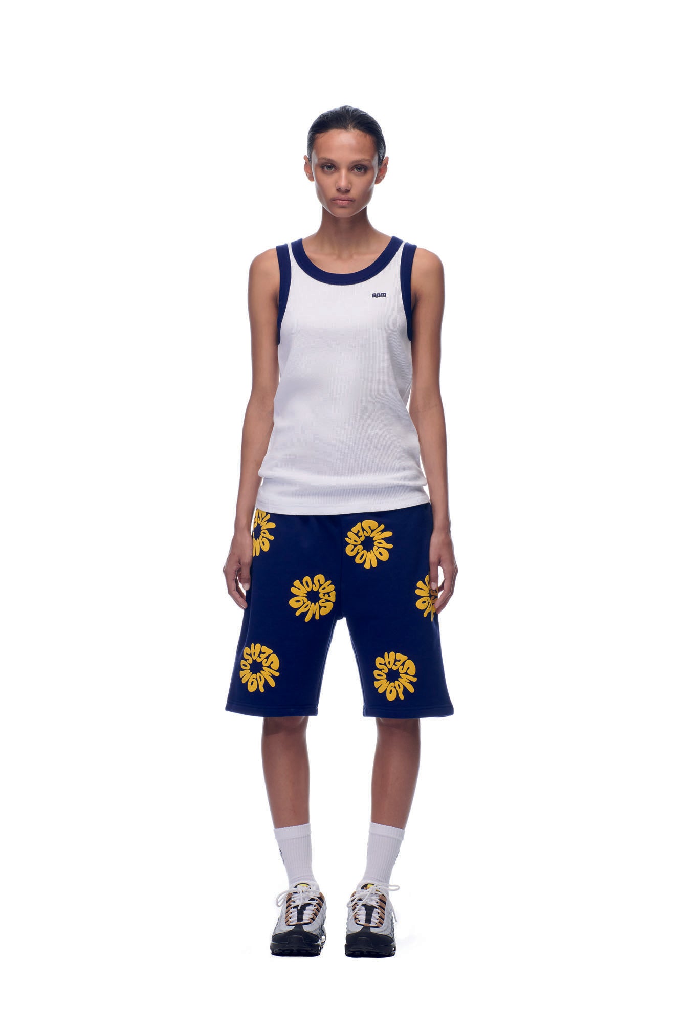 A person faces forward against a plain white background, wearing the 6PM NH TANKTOP WHITE/NAVY, blue knee-length shorts with yellow flower patterns, white socks, and sneakers.