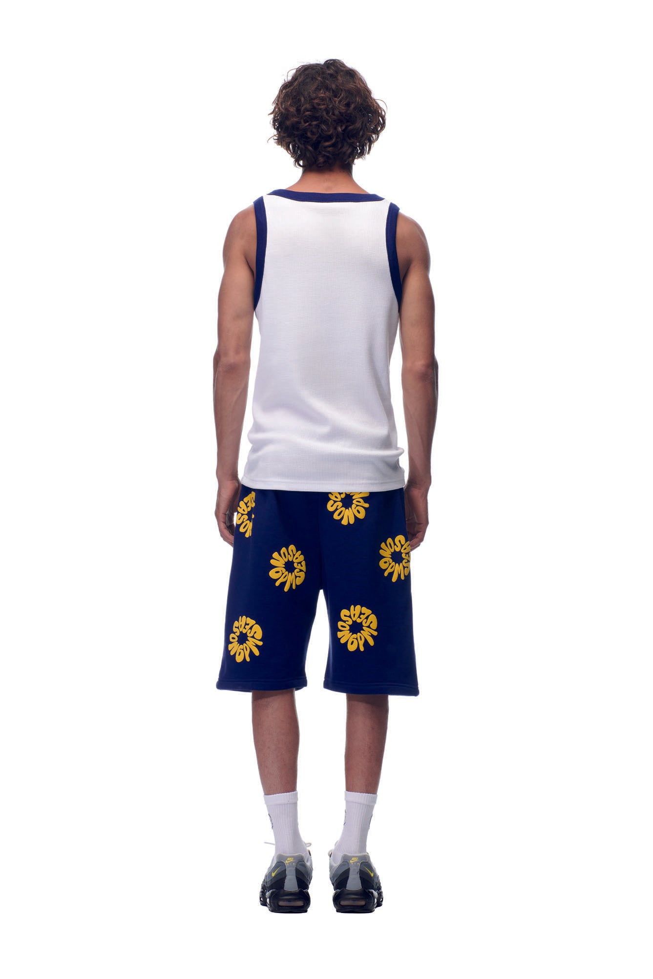 A person faces away, wearing the 6PM NH TANKTOP WHITE/NAVY with blue knee-length shorts featuring yellow floral patterns, white socks, and black sneakers, set against a plain white background.
