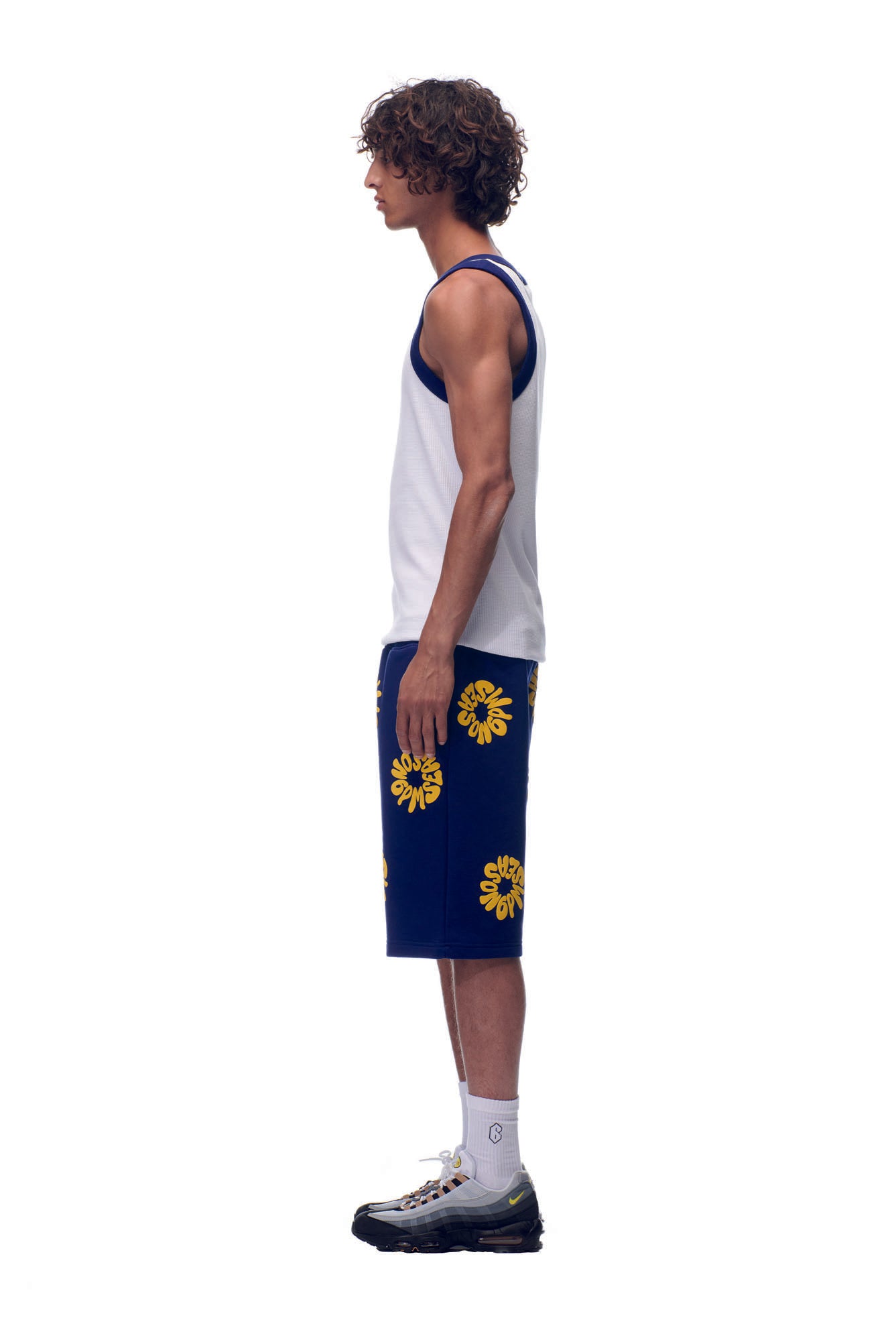 A young man stands in profile against a white background, wearing the 6PM NH TANKTOP WHITE/NAVY, navy blue shorts with yellow floral patterns, white socks, and black sneakers.